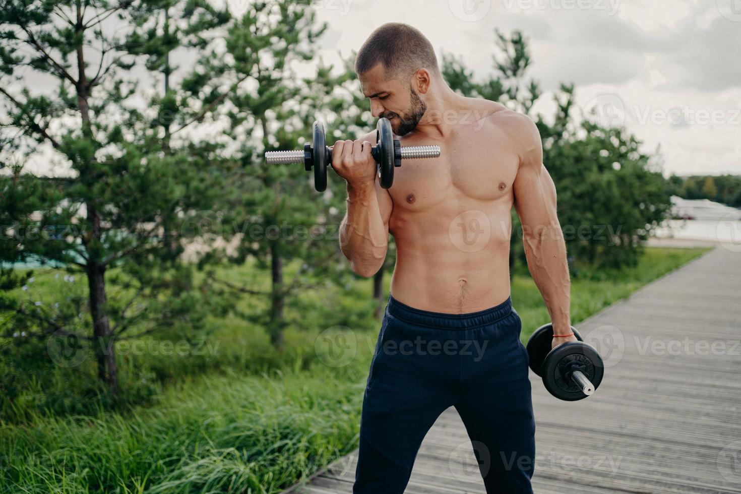 tiro horizontal de un hombre musculoso motivado que trabaja con una barra demuestra el poder de la masculinidad tiene un cuerpo fuerte se para sin camisa al aire libre se esfuerza por levantar peso con esfuerzo hace rizos de bíceps foto