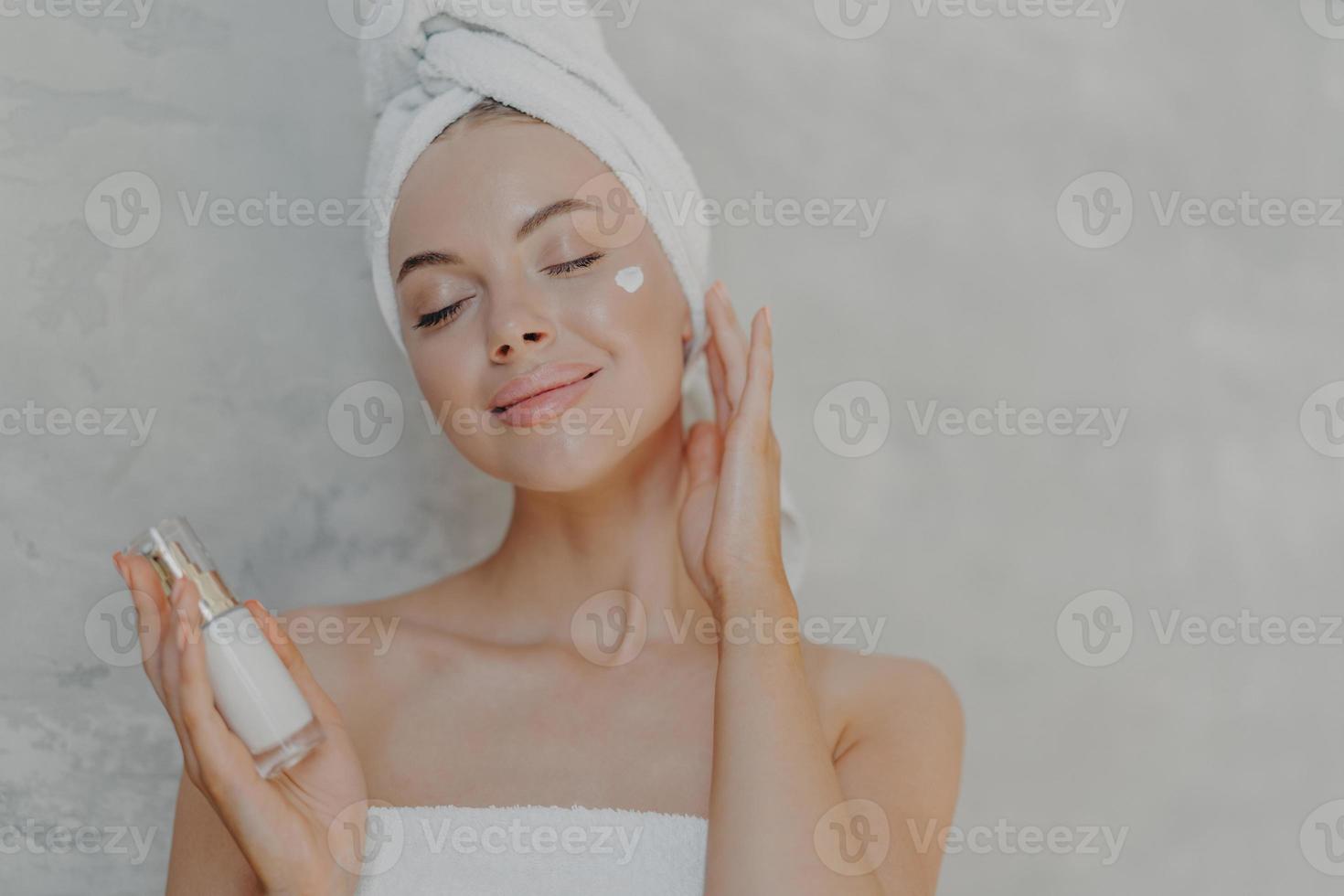 Headshot of pleased attractive woman applies face lotion, satisfied with new cosmetic product, keeps eyes closed, touches soft skin after bath, has well cared complexion, poses against grey wall photo