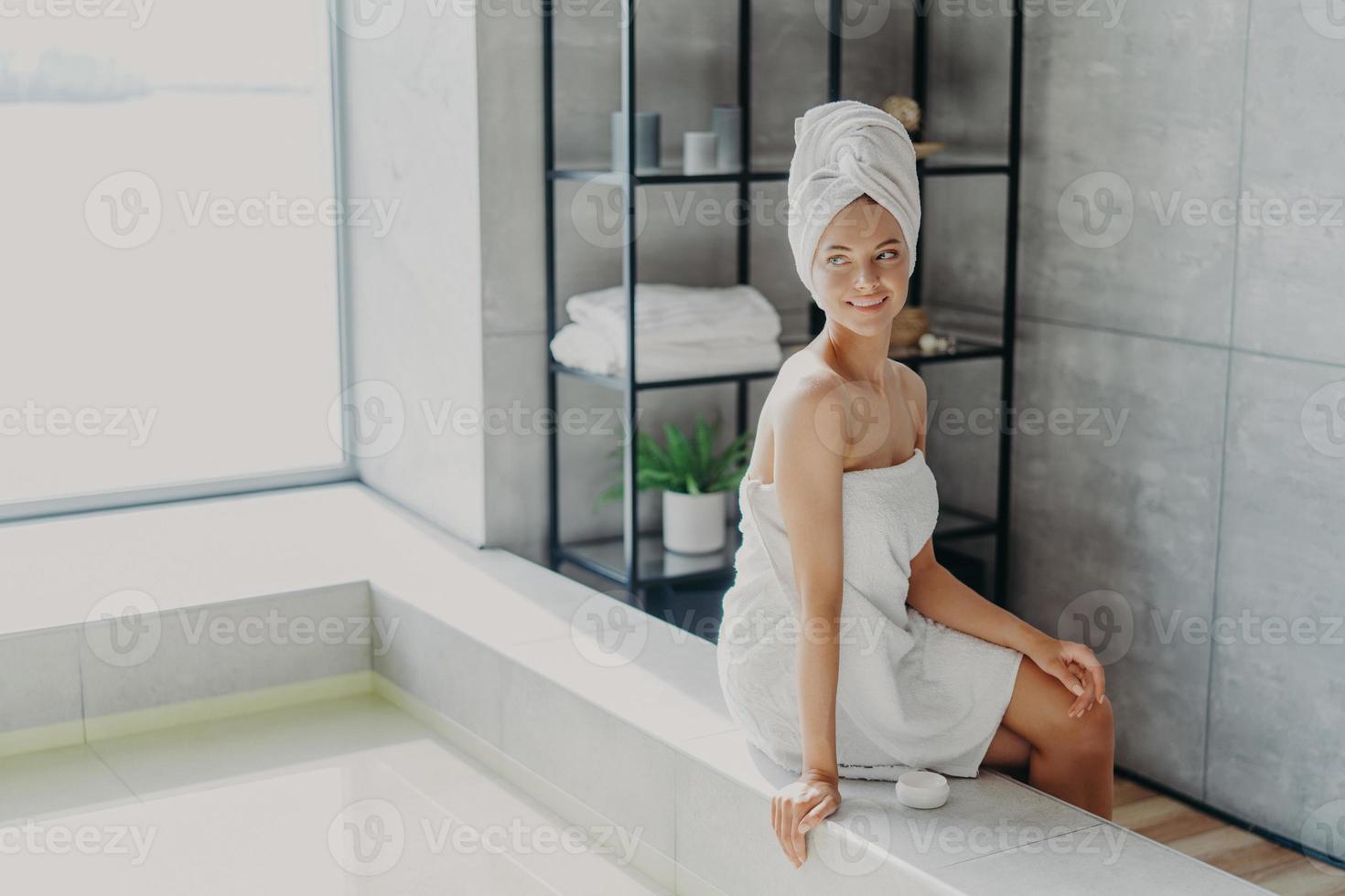 Beauty model with healthy perfect skin applies cream for body care, poses wrapped in towel after taking shower, poses at bathtube, looks thoughtfully away, feels refreshed and relaxed. Spa procedures photo