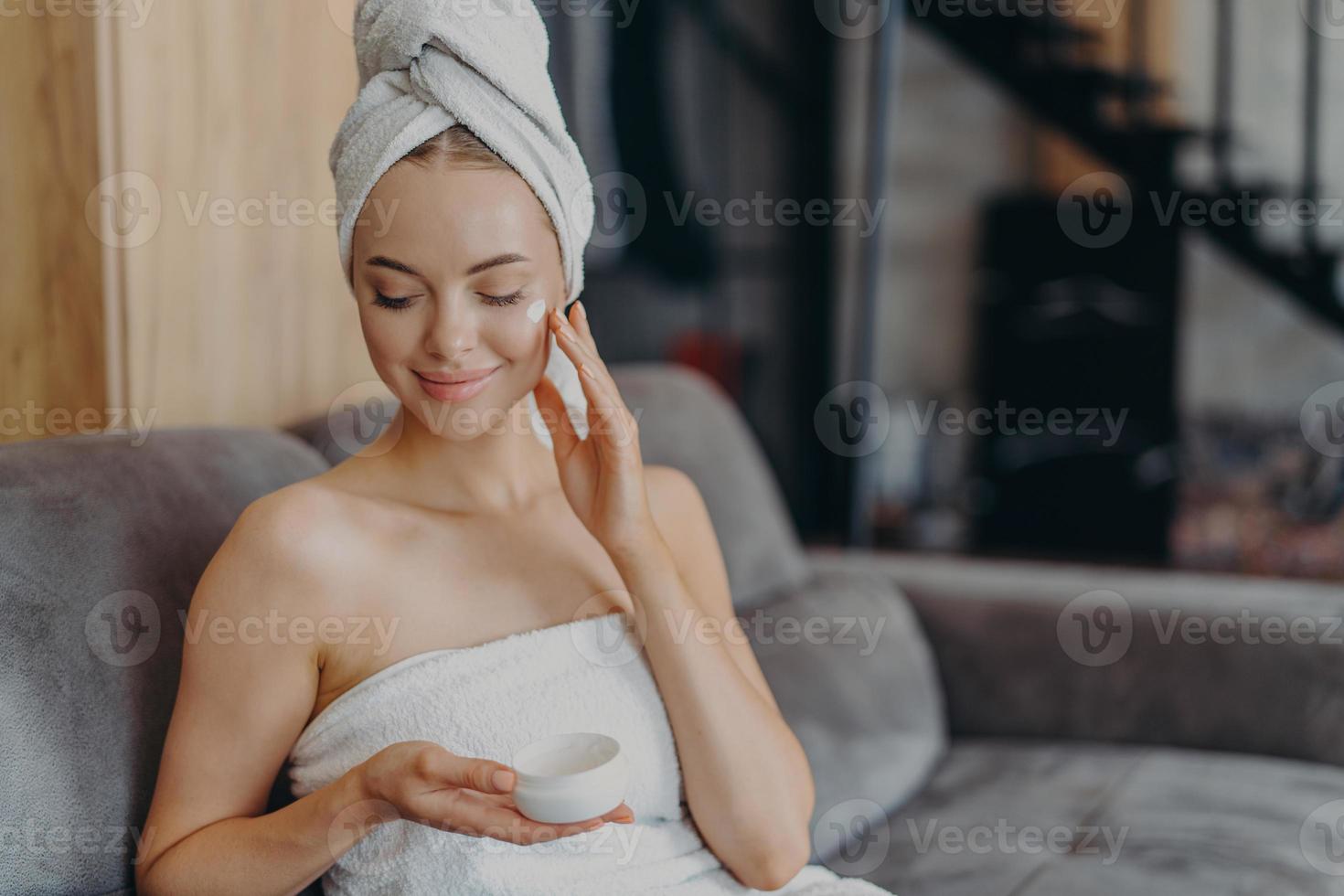 Beautiful female model enjoys softness of skin afrer taking bath, applies moisturizing cream on cheek, wears minimal makeup, wrapped in towel, sits on sofa alone, undergoes beauty treatments photo
