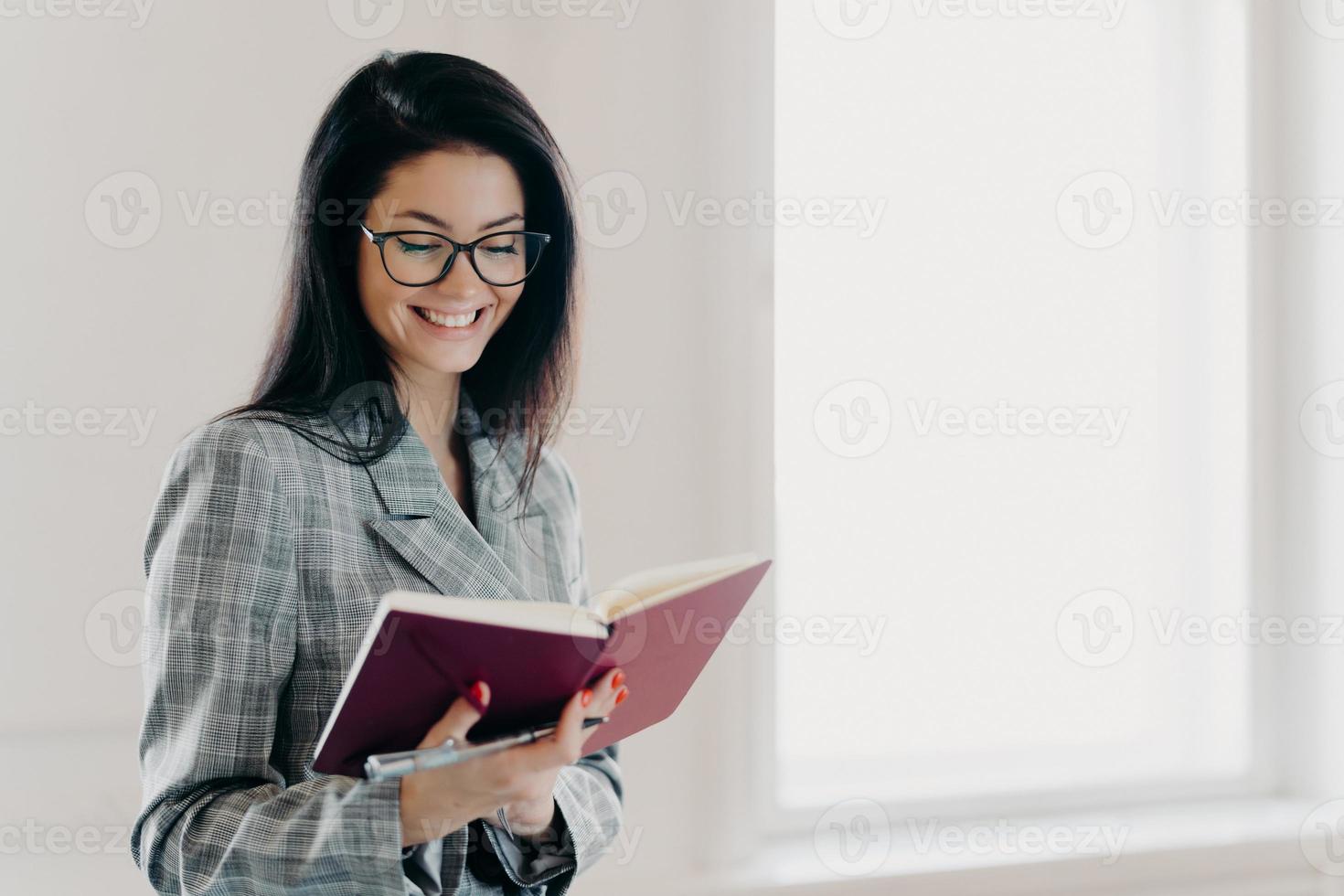 mujer morena satisfecha tiene expresión feliz anota el plan de trabajo en el bloc de notas tiene manicura roja ropa formal registra información para el proceso de organización usa anteojos notas calendario financiero foto