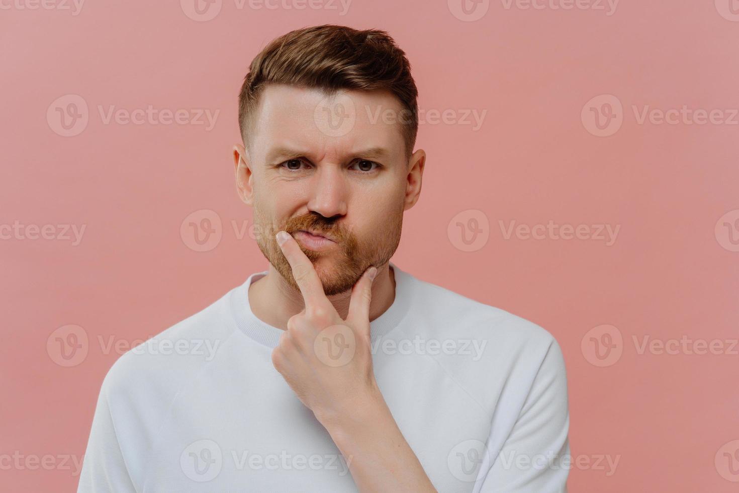 Photo of serious man holds chin purses lips makes decision with doubtful expression focused displeased at camera dressed in casual white t shirt isolated over pink background. Let me thinks a bit