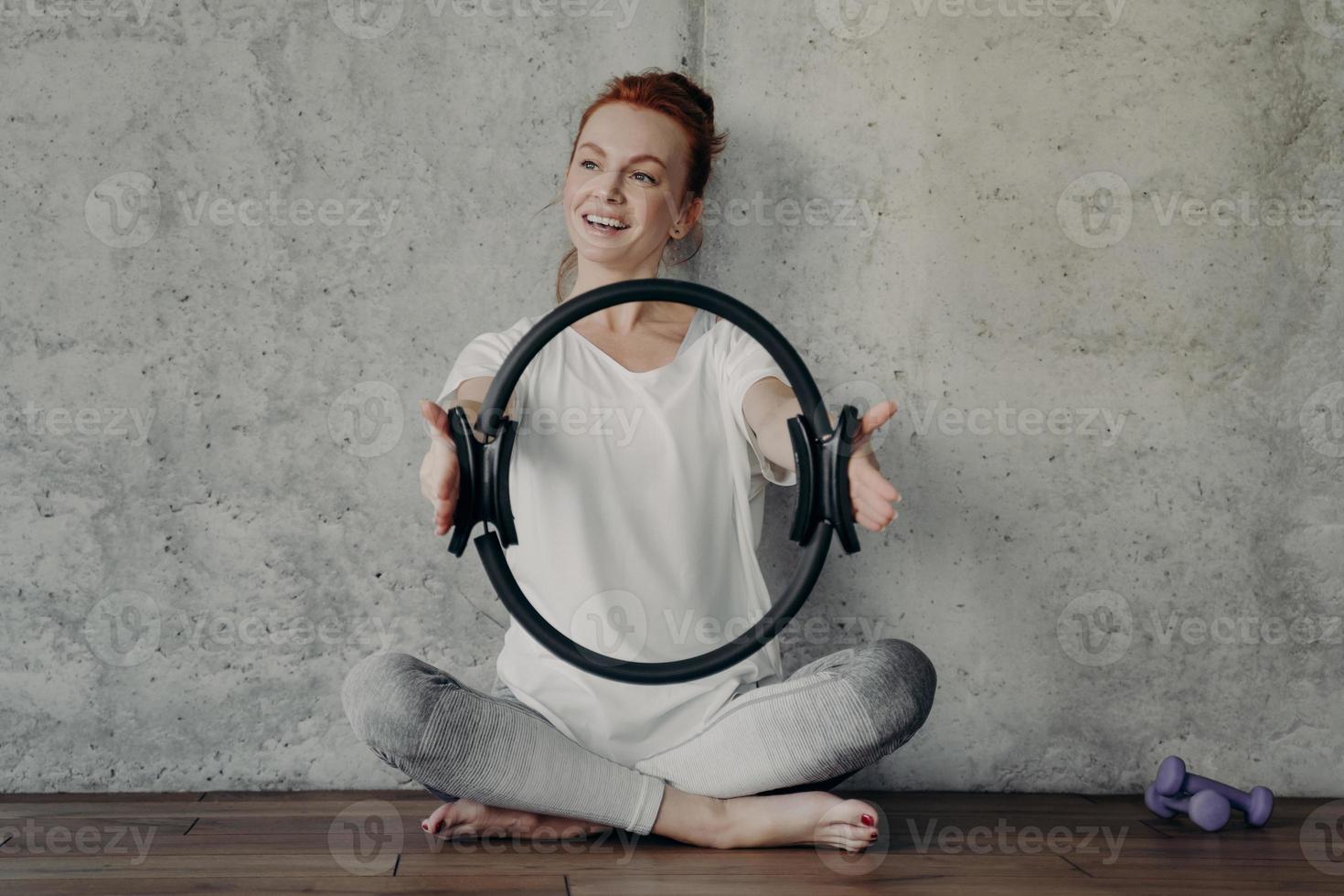 feliz mujer pelirroja sonriente sentada en posición de loto con anillo de pilates durante el entrenamiento foto
