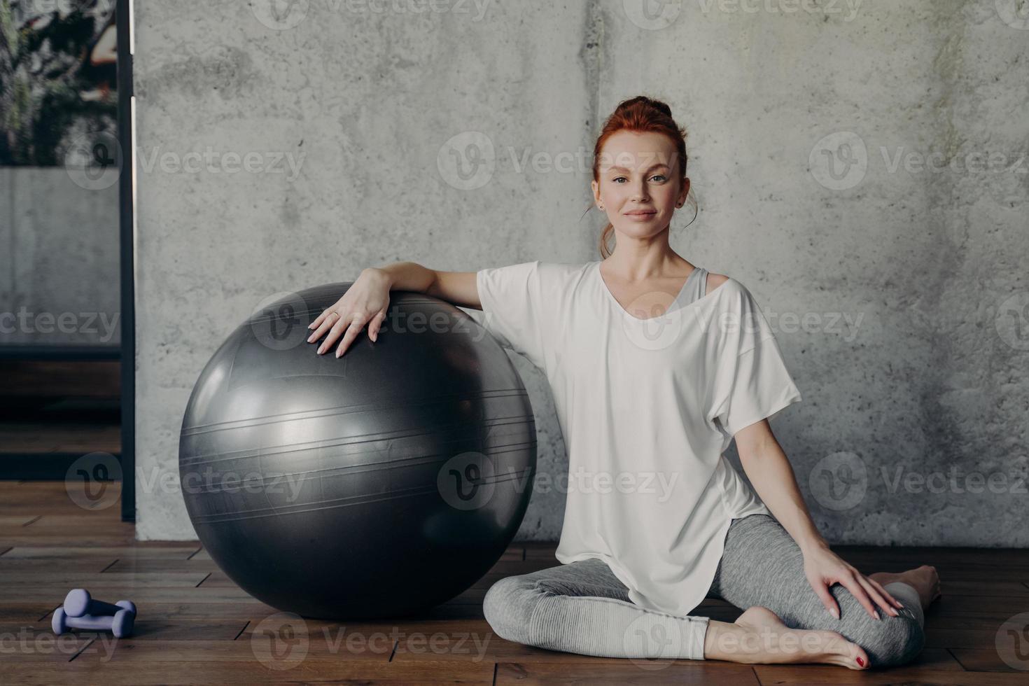 Beautiful sportive red haired lady gracefully sits on parquet floor with fitball photo