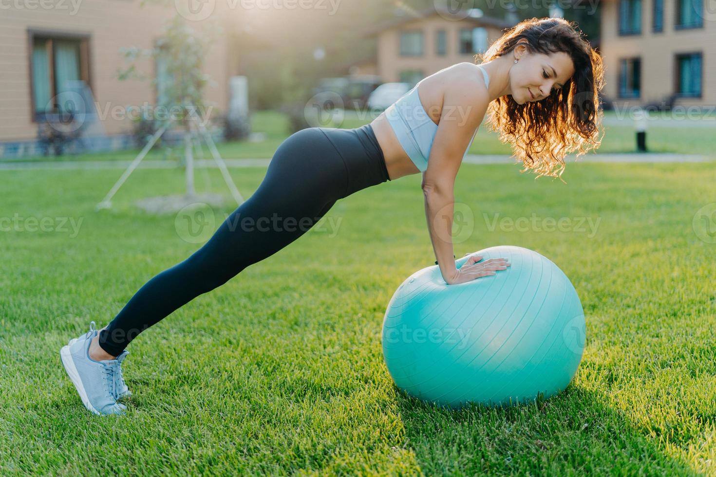 Active fitness woman does gymnastic outdoor with inflated fit ball, has active workout, makes physical exercises in open air, dressed in sport clothes trains on green grass. Active lifestyle concept photo