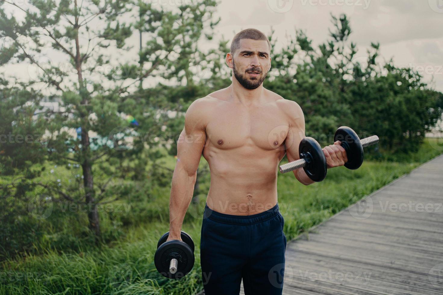 Handsome muscular man lifts barbells outside, does biceps training, stands with naked sexy torso, trains muscles, dressed in shorts poses outdoor, has attractive body. Weight lifting concept photo