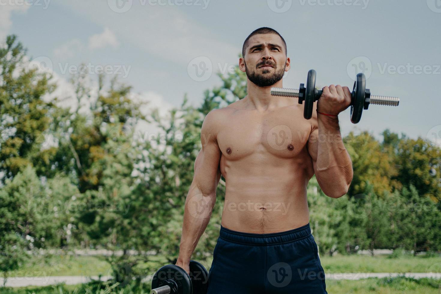 Muscular shirtless young man raises barbells outdoors, trains muscles and has strong body. Athletic sportsman with strong arms leads healthy lifestyle, enjoys training against nature background. photo