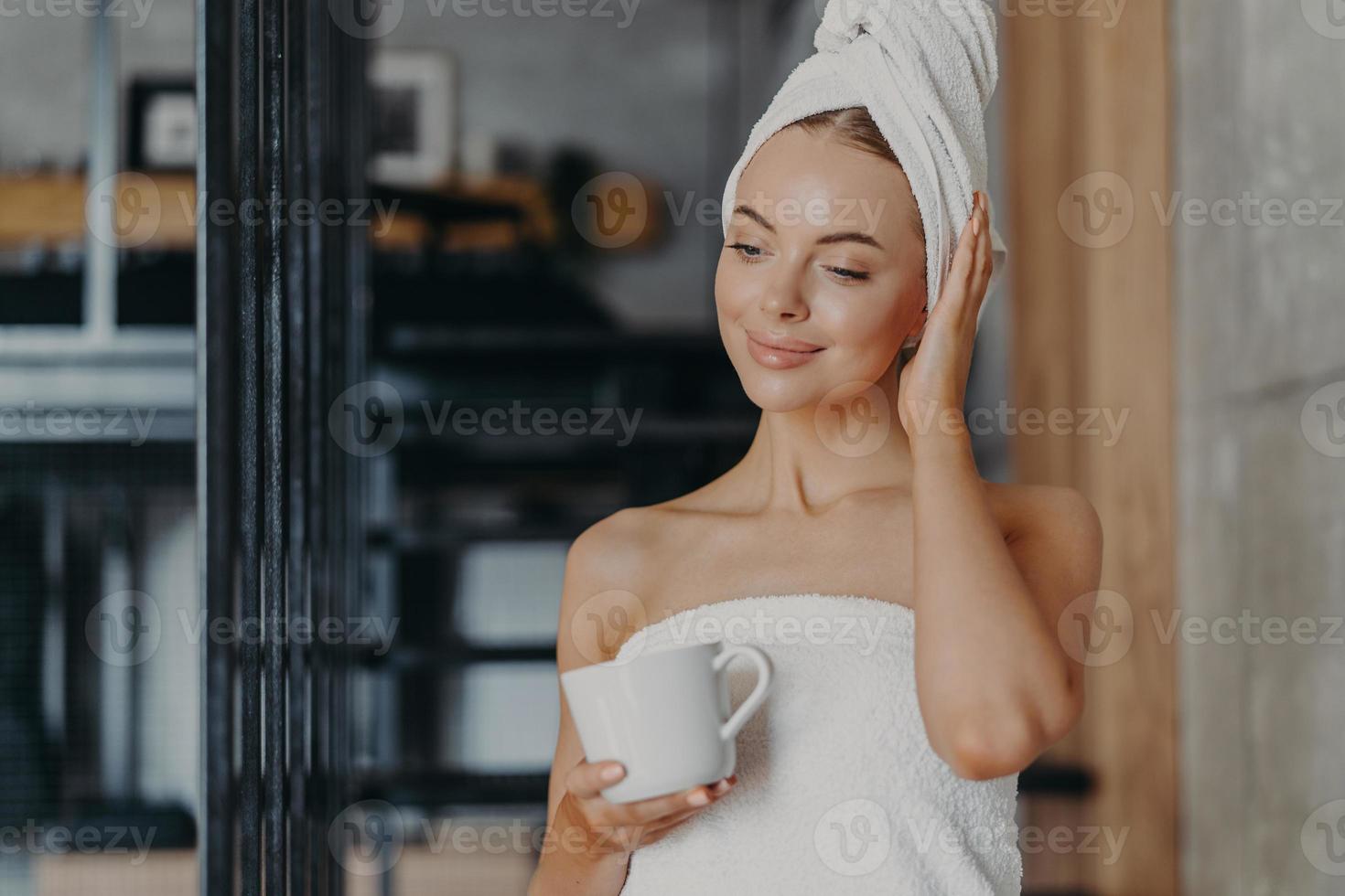Photo of healthy thoughtful woman with healthy smooth skin drinks hot tea concentrated down, stands wrapped in white bath towel in cozy room, enjoys hygiene and beauty treatments, feels relaxed
