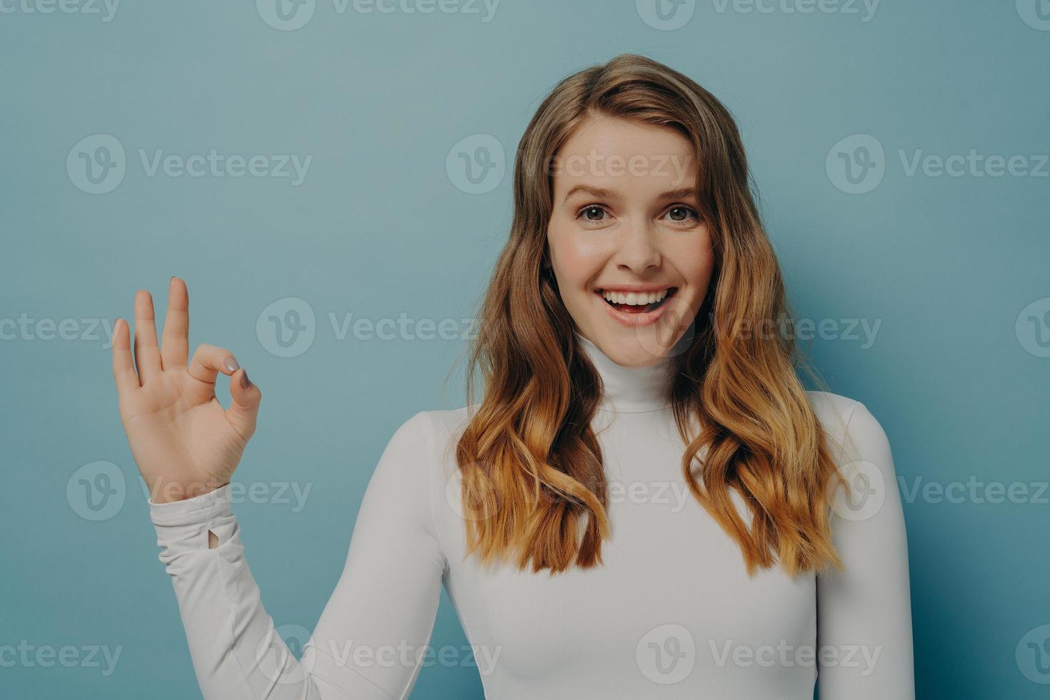 Beautiful excited joyful young lady with dyed light-brown hair gesturing ok symbol with fingers photo
