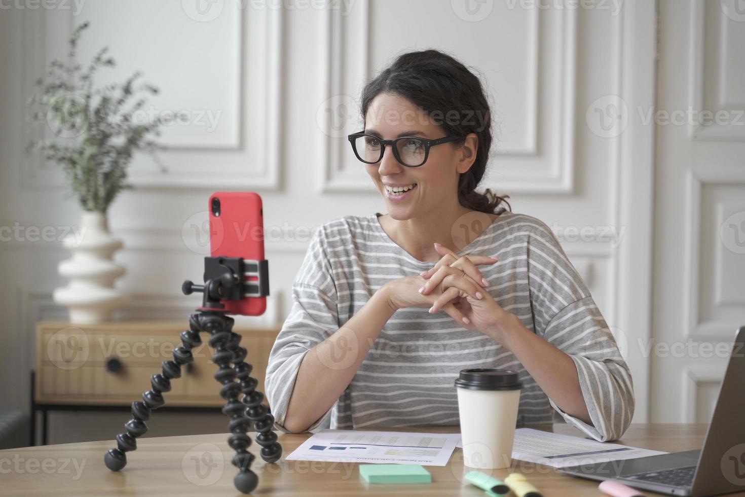joven empresaria española sonriente con gafas sentada frente a un smartphone en un trípode en el lugar de trabajo foto