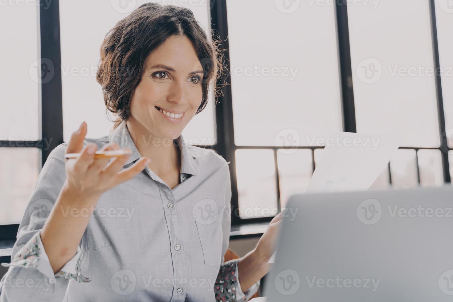 Cheerful woman Spanish tutor looks at laptop screen with confident smile while explaining new topic photo