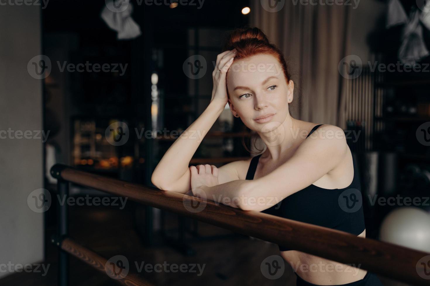 joven mujer atractiva con el pelo rojo se apoya en la barra de ballet y descansa después del entrenamiento foto