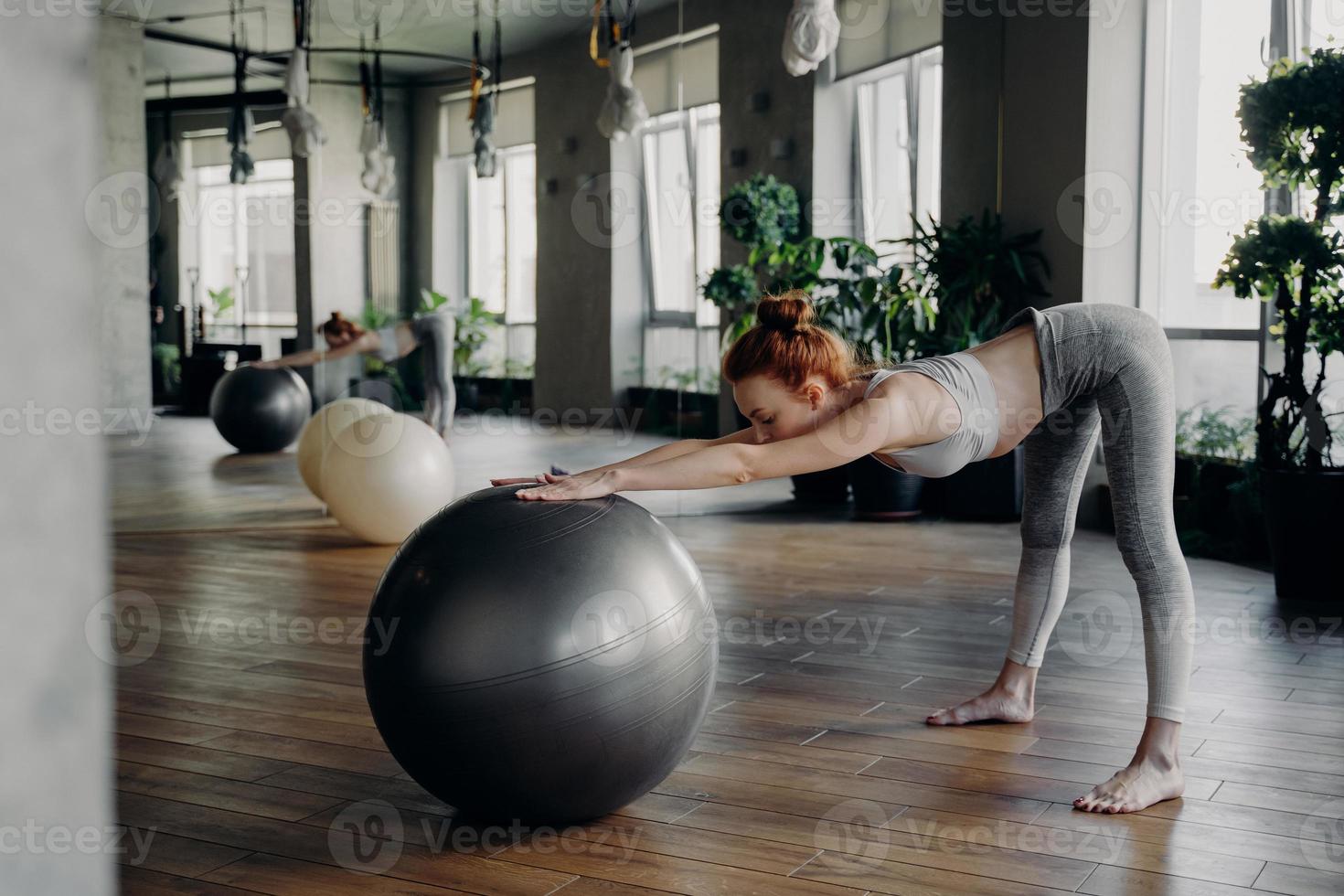 Slim woman stretching back with exercise ball while working out in modern fitness studio photo