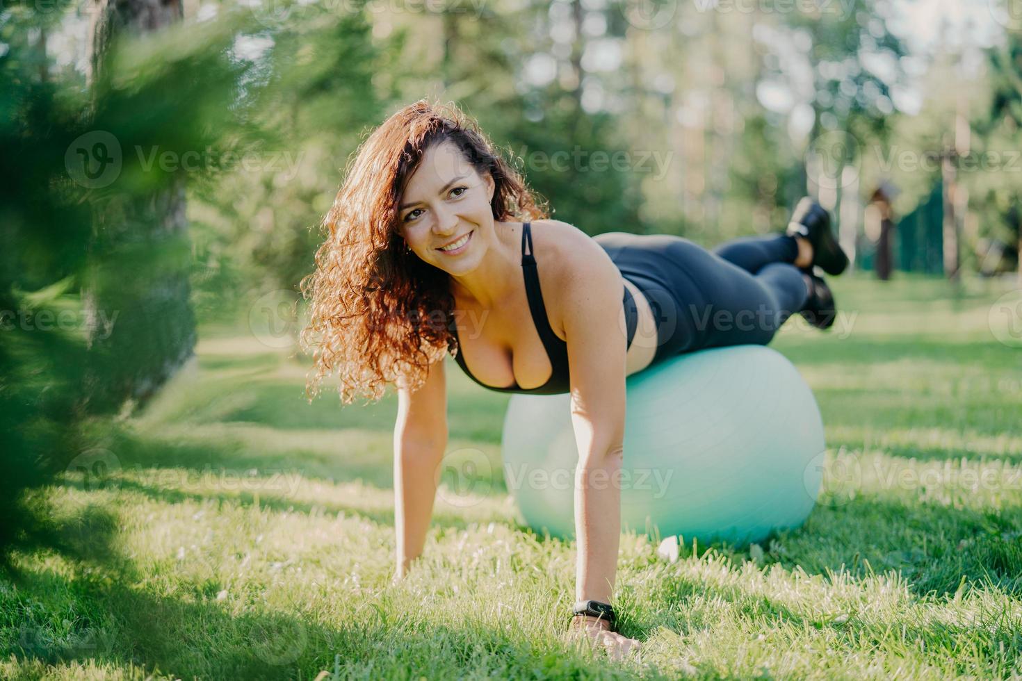 Happy active sportswoman does fitness exercises with gymnastic ball, dressed in active wear, has workout in open air, poses on green grass in forest or park, focused somewhere with pleasant smile photo