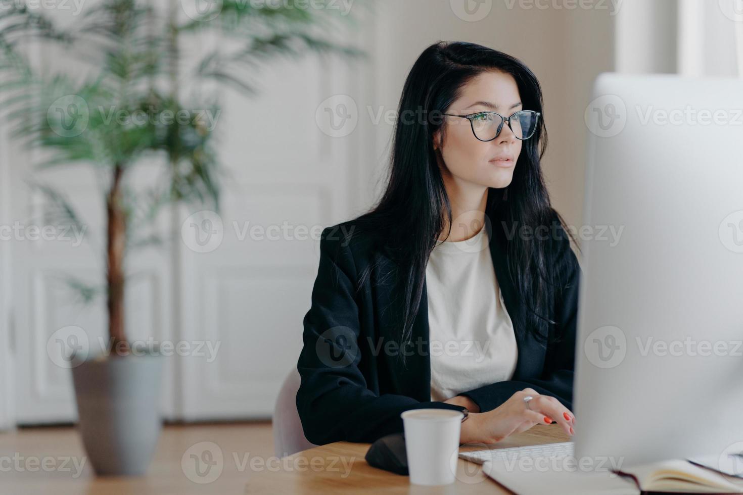 Elegant brunette businesswoman poses at cowroking space, concentrated at monitor of computer, dressed formally, drinks aromatic coffee, analyzes information, works in office, has serious expression photo