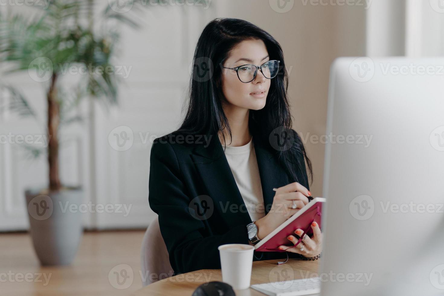 Indoor shot of prosperous businesswoman makes notes in notepad, concentrated in computer screen, watches training video how to start own business, poses at desktop with coffee cup, makes report photo