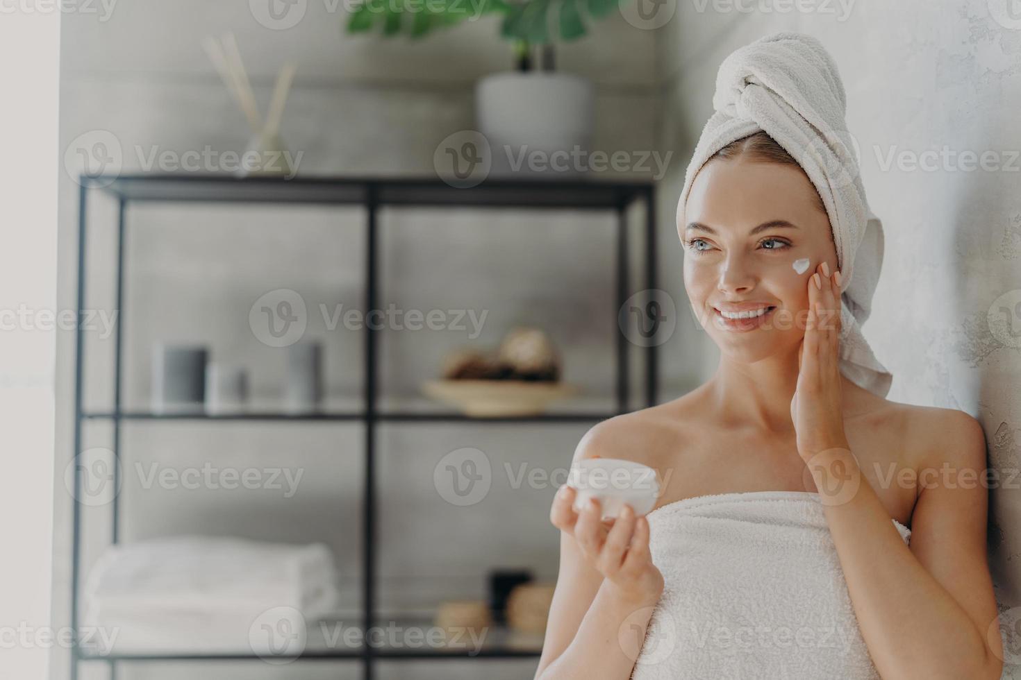 foto de una mujer europea muy joven que aplica crema hidratante en la cara, tiene una expresión feliz, envuelta en una toalla de baño, se alza contra el fondo del baño interior. concepto de higiene y cuidado de la piel