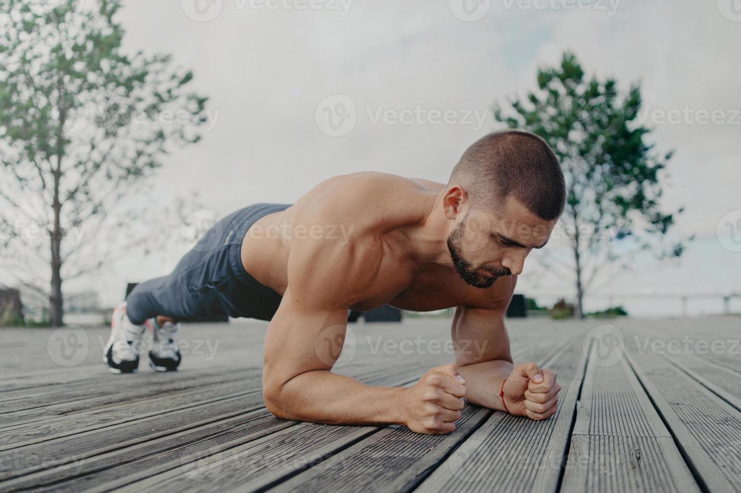 tiro al aire libre de deportista fuerte se encuentra en pose de tablón, pone todos los esfuerzos para mantenerse en forma y saludable. hombre atleta con expresión concentrada, trabaja al aire libre. culturista hace ejercicio de prensa foto