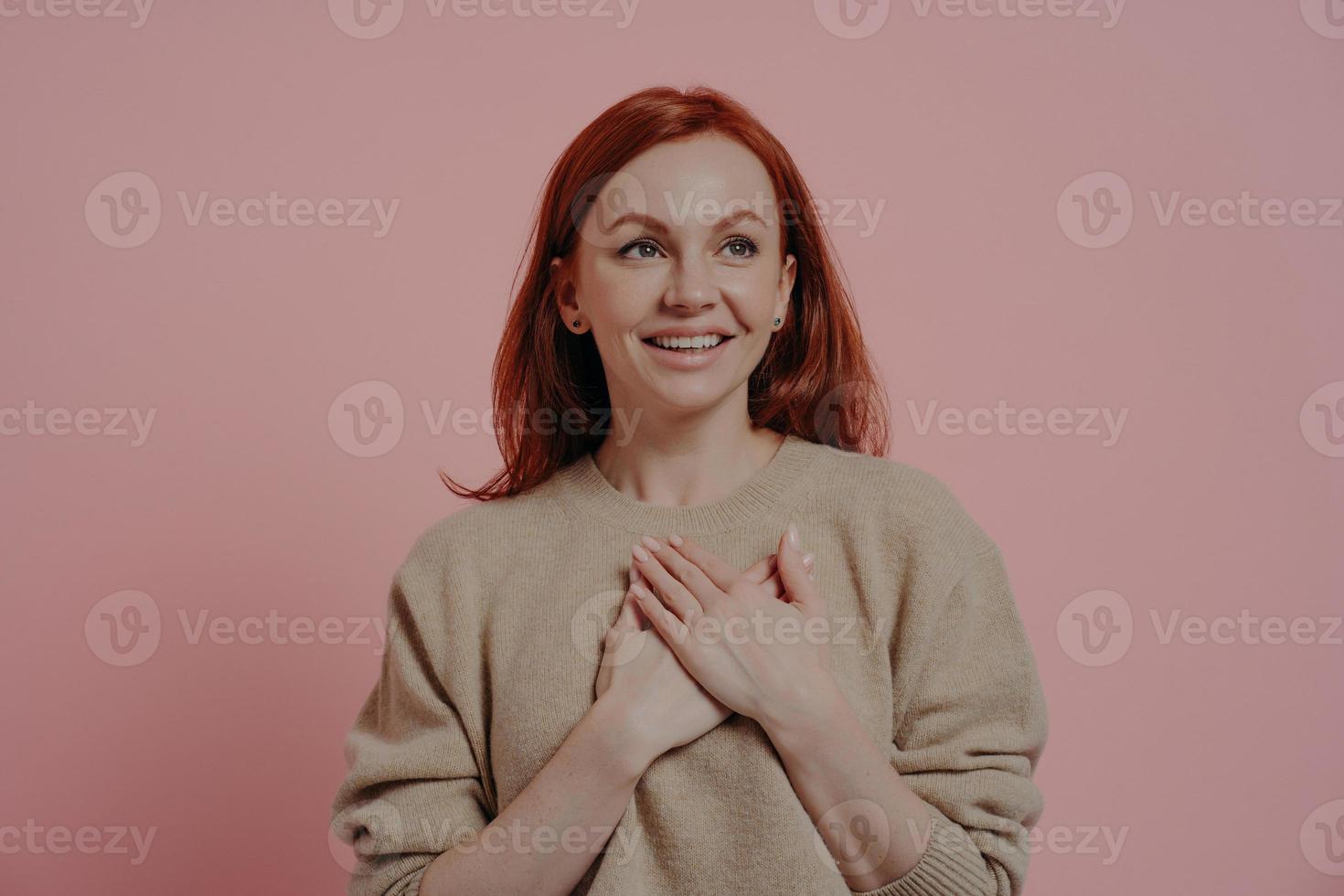 Young happy red-haired woman putting hands on chest while standing isolated on pink background photo