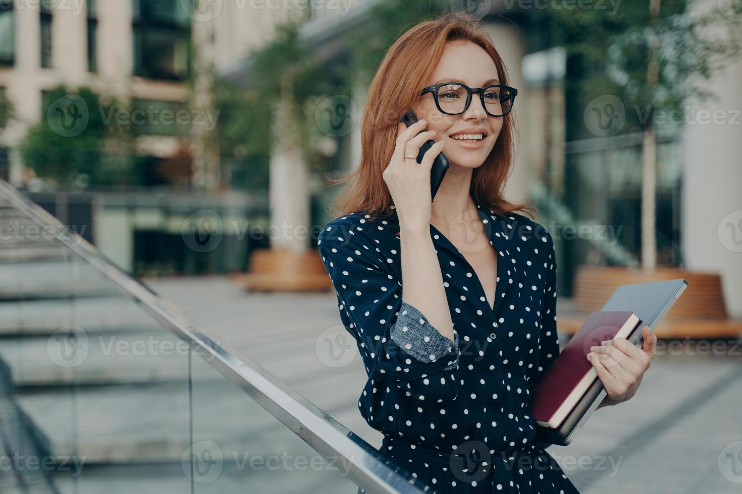 Positive young redhead woman enjoys mobile phoning keeps smartphone near ear photo
