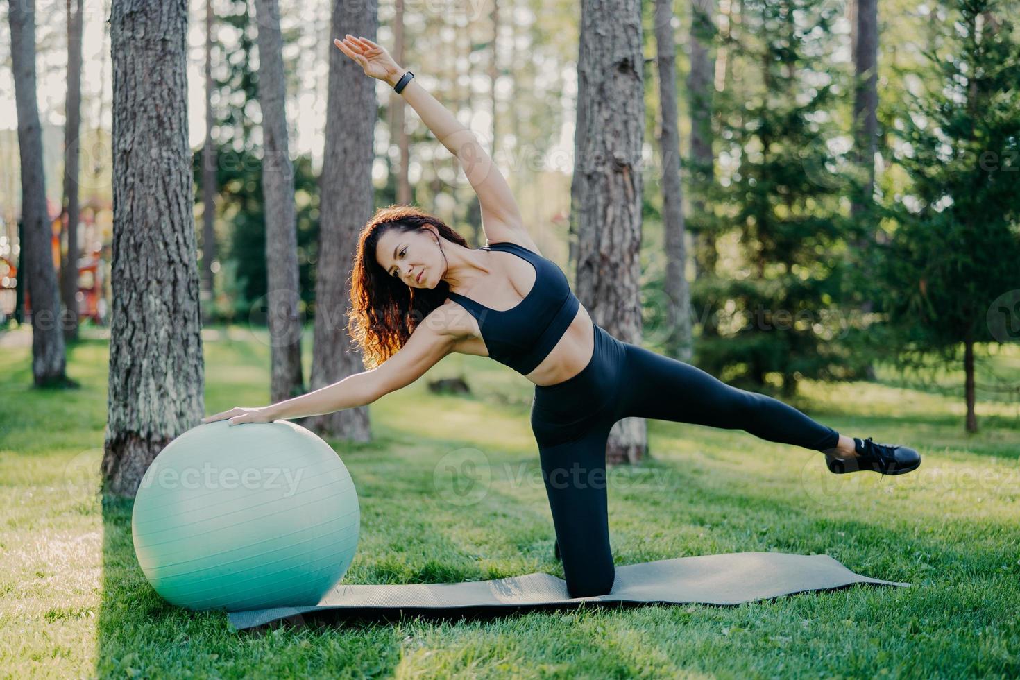 foto al aire libre de una mujer morena activa en poses de ropa deportiva en una alfombra de yoga, hace ejercicios de estiramiento con pelota gimnástica, posa en el bosque o parque sobre hierba verde. aeróbic, concepto de estilo de vida saludable