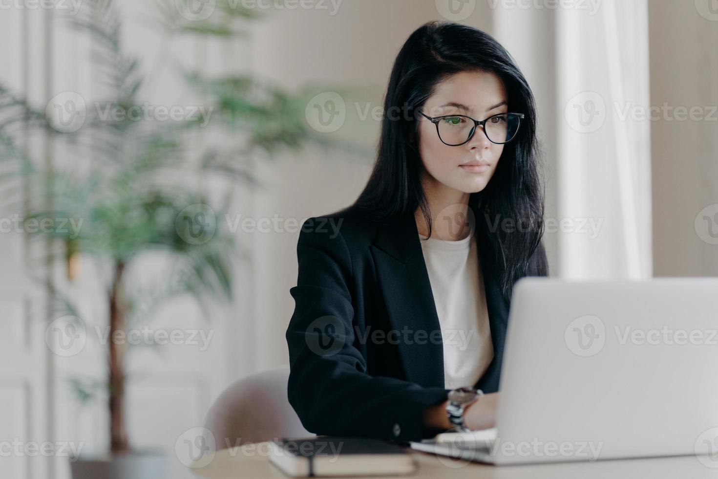 Young serious businesswoman works with laptop computer at workplace in office, wears optical glasses and formal costume, concentrated at screen. Job, occupation, business development concept photo