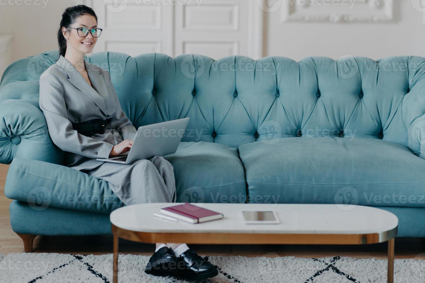 Young prosperous female executive surfs on official site of media marketing corporation uses modern laptop computer, uses wireless internet, works in cozy cabinet sits on comfortable sofa with desktop photo