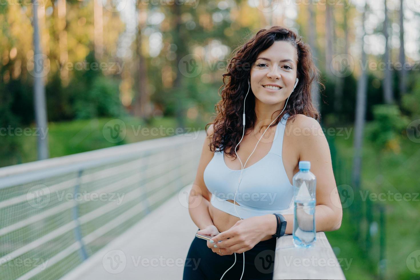 Photo of slim beautiful curly woman dressed in cropped top and leggings enjoys listening music holds mobile phone drinks fresh water from bottle poses at bridge poses against nature background