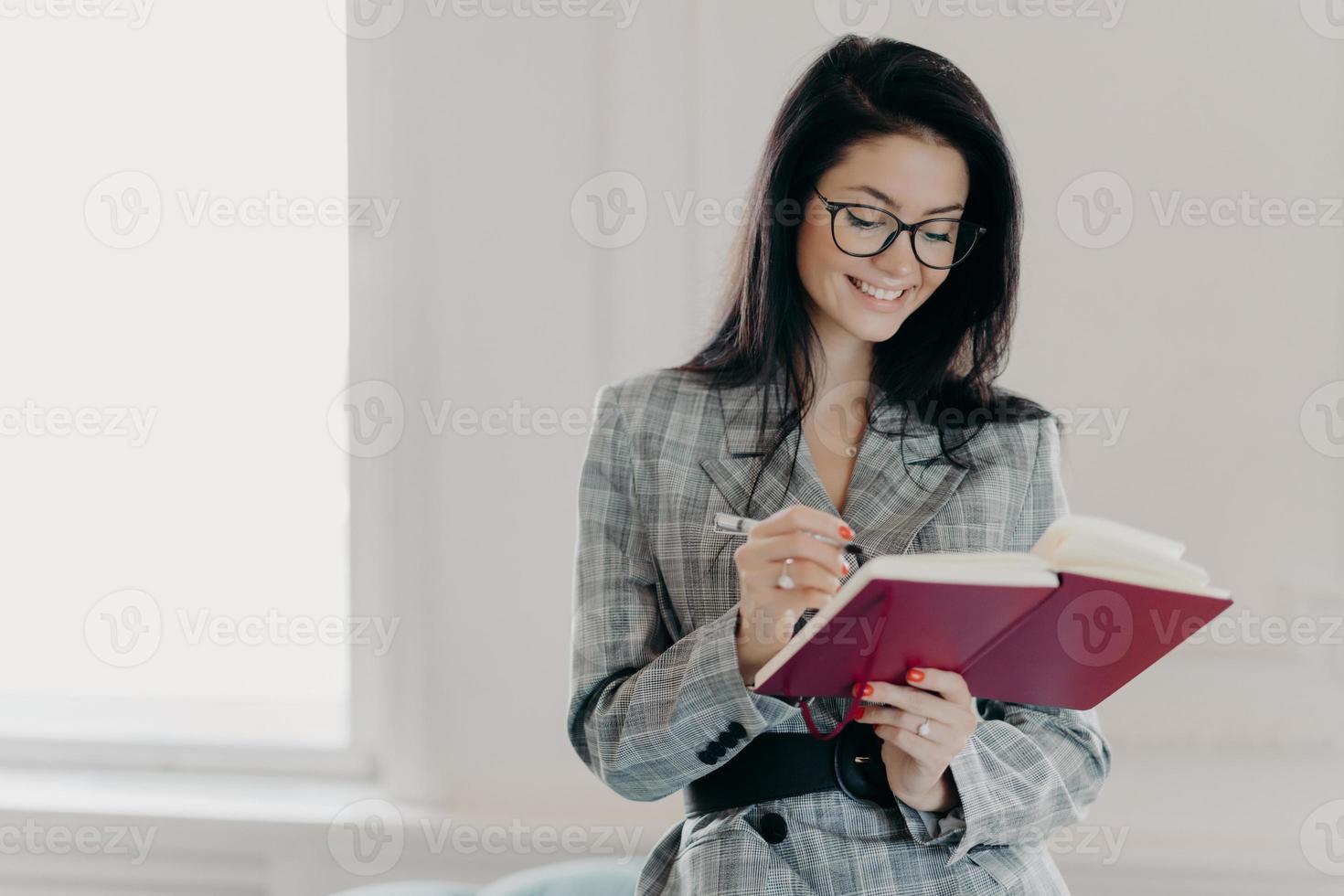 Horizontal shot of good looking woman professor writes down text notes in notepad, prepares for lecture in university, wears transparent glasses, business suit, stands indoor with broad smile photo