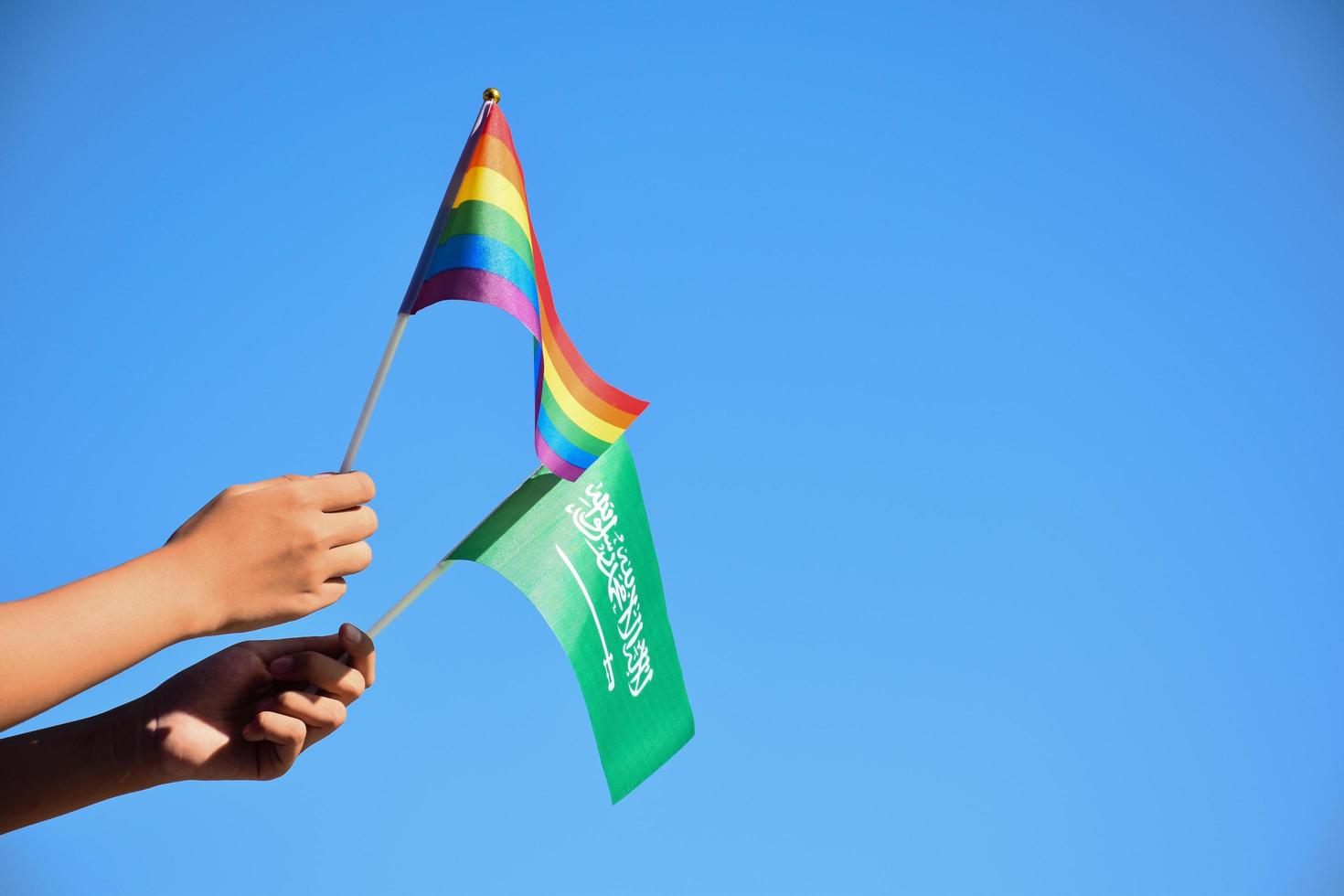 Saudi Arabia flag and rainbow flag, LGBT symbol, holding in hands, blue sky background, concept for LGBT celebration in Saudi Arabia in pride month, June, soft and selective focus. photo