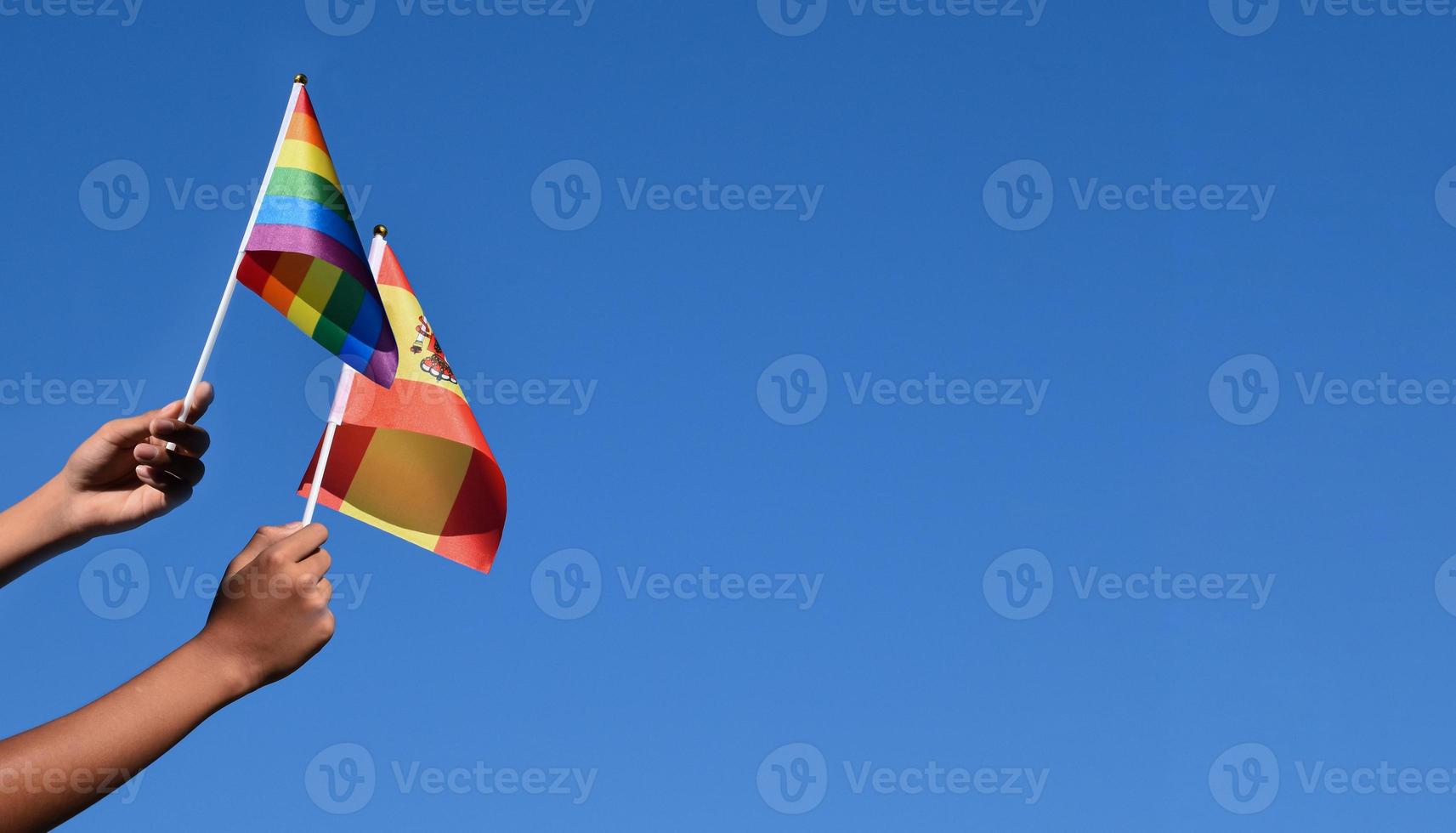 Spain flag and rainbow flag, LGBT symbol, holding in hands, bluesky background, concept for LGBT celebration in Spain and around the world in pride month, June, soft and selective focus, copy space. photo