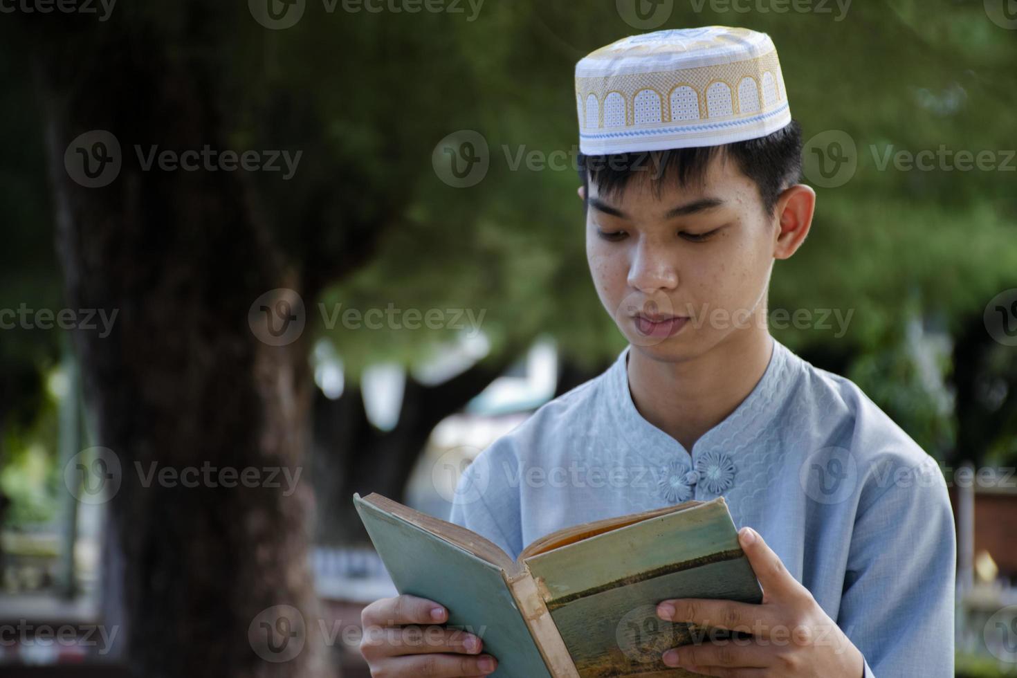 Young asian muslim boy sitting in school park and reading his book in his free times before going back home, soft and selective focus. photo