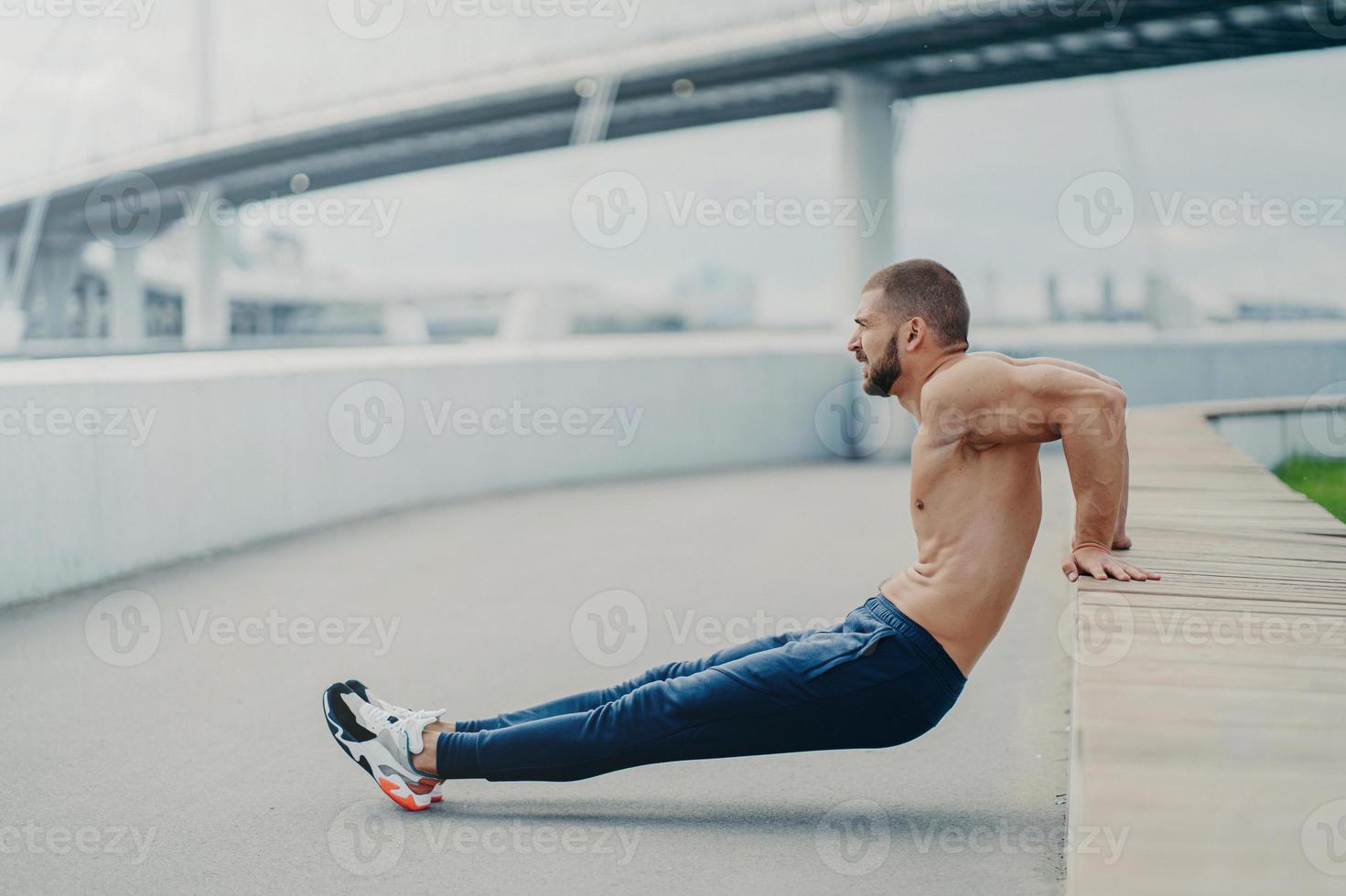 Sideways shot of muscular man does reverse push up exercise, trains arms, keeps perfect body and good physical shape, has active workout outdoor, wears sport trousers and sneakers, poses on bridge photo