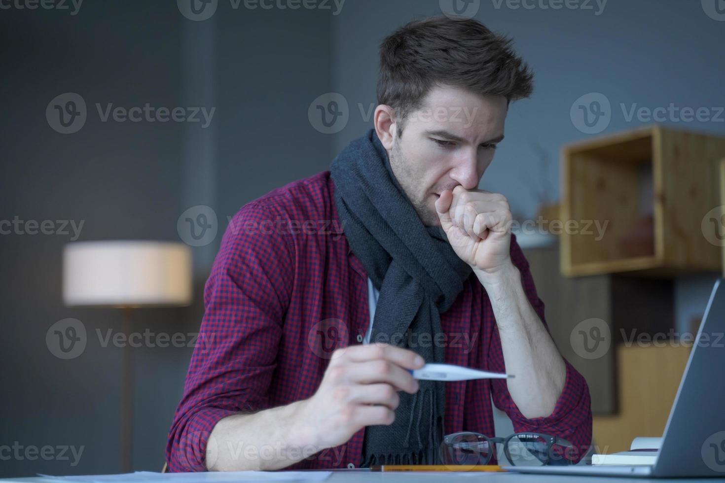 Unhealthy looking male entrepreneur in scarf around his neck coughing looks at digital thermometer photo