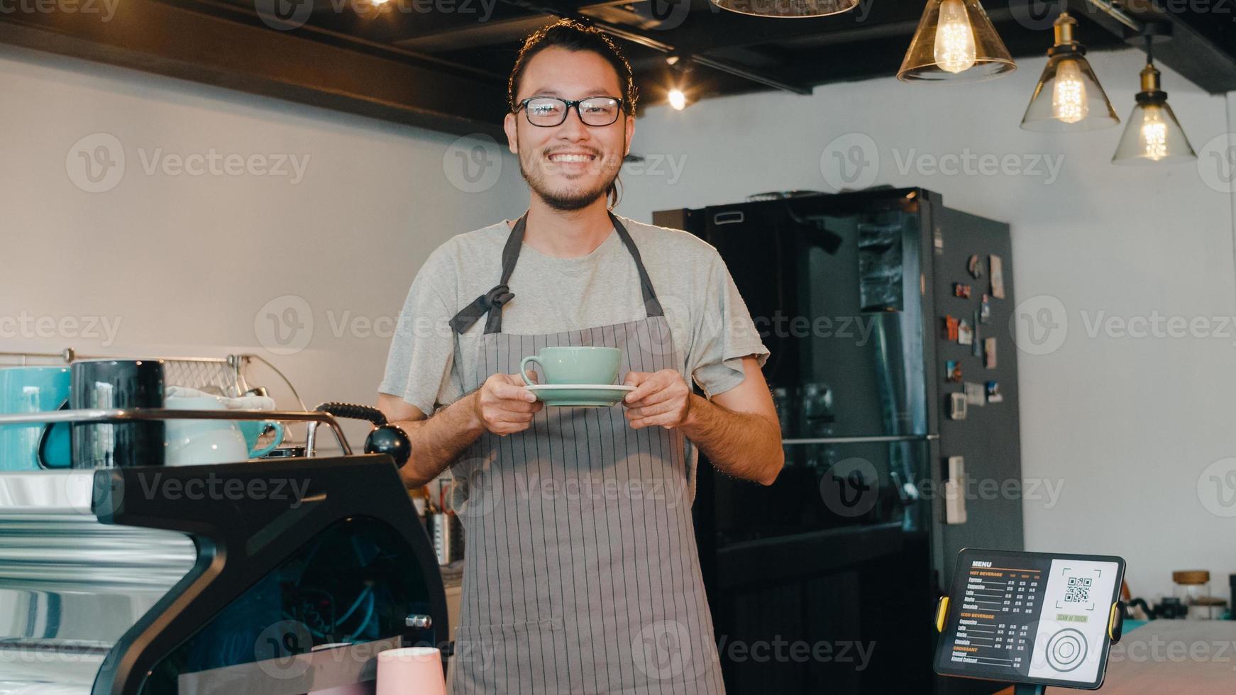 Portrait young Asian guy barista waitress holding coffee cup feeling happy at urban cafe. Asia small business owner boy in apron relax toothy smile looking to camera stand at counter in coffee shop. photo