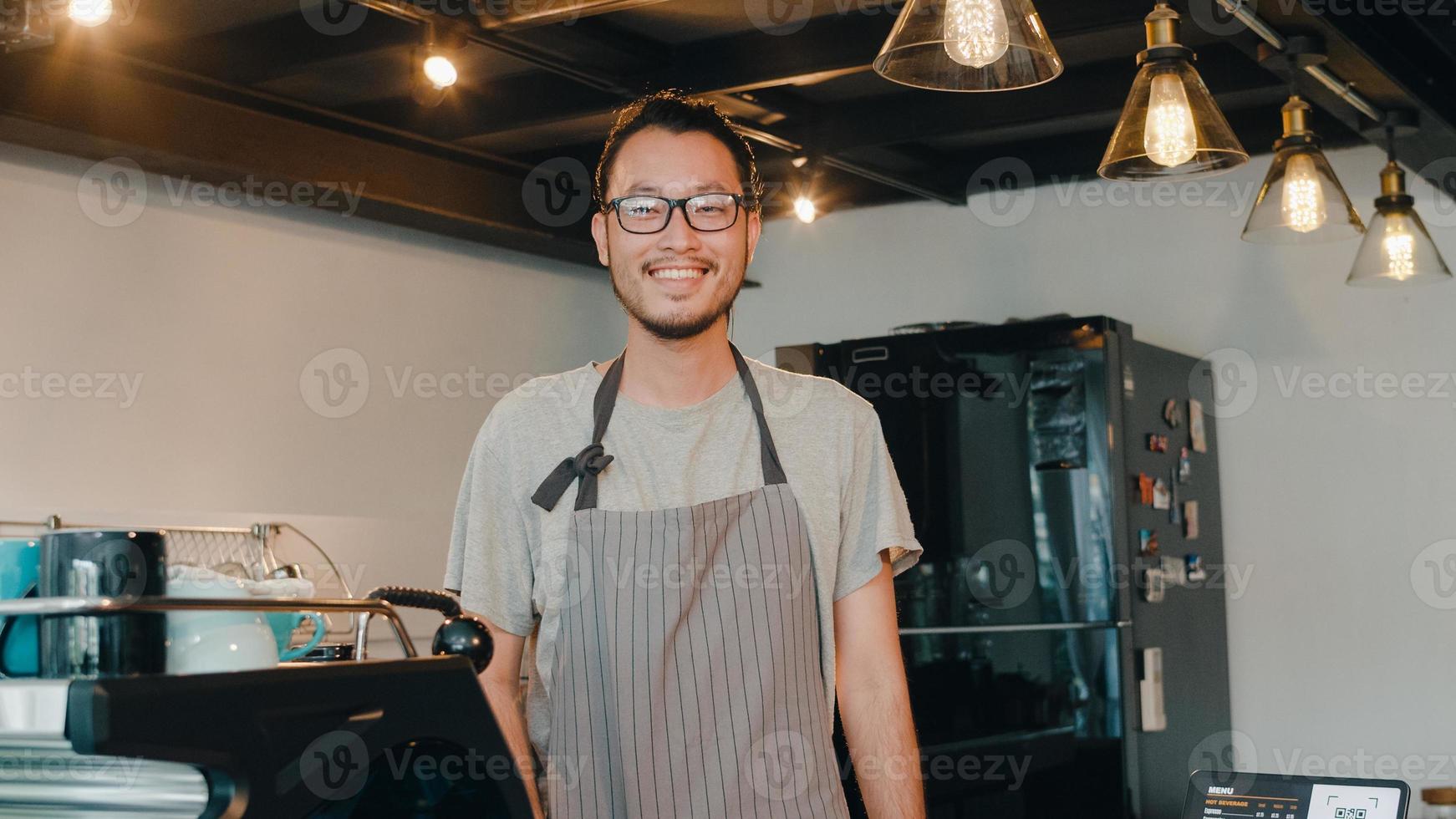 Portrait young Asian man barista feeling happy smiling at urban cafe. Small business owner Japanese male in apron relax toothy smile looking to camera standing at the counter in coffee shop. photo