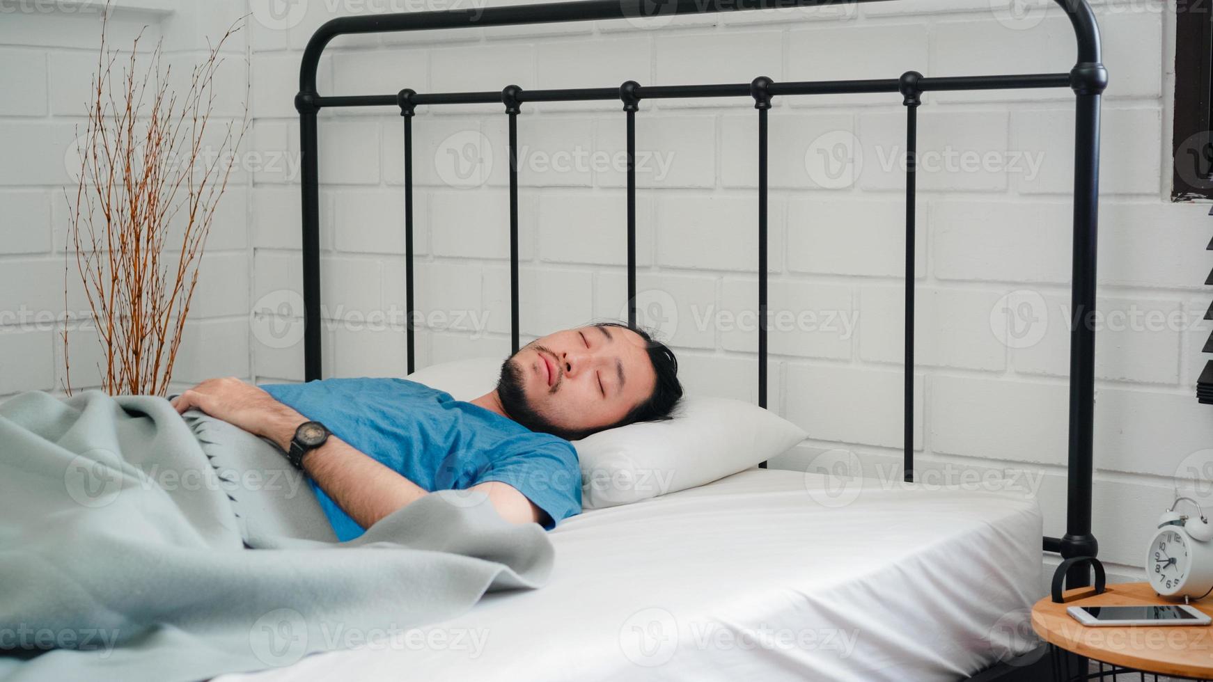 un joven asiático se despierta por la mañana, un hombre se estira después de despertarse en la cama en el dormitorio de su casa. los hombres guapos toman una siesta, se relajan con sueño en el concepto de casa moderna. foto