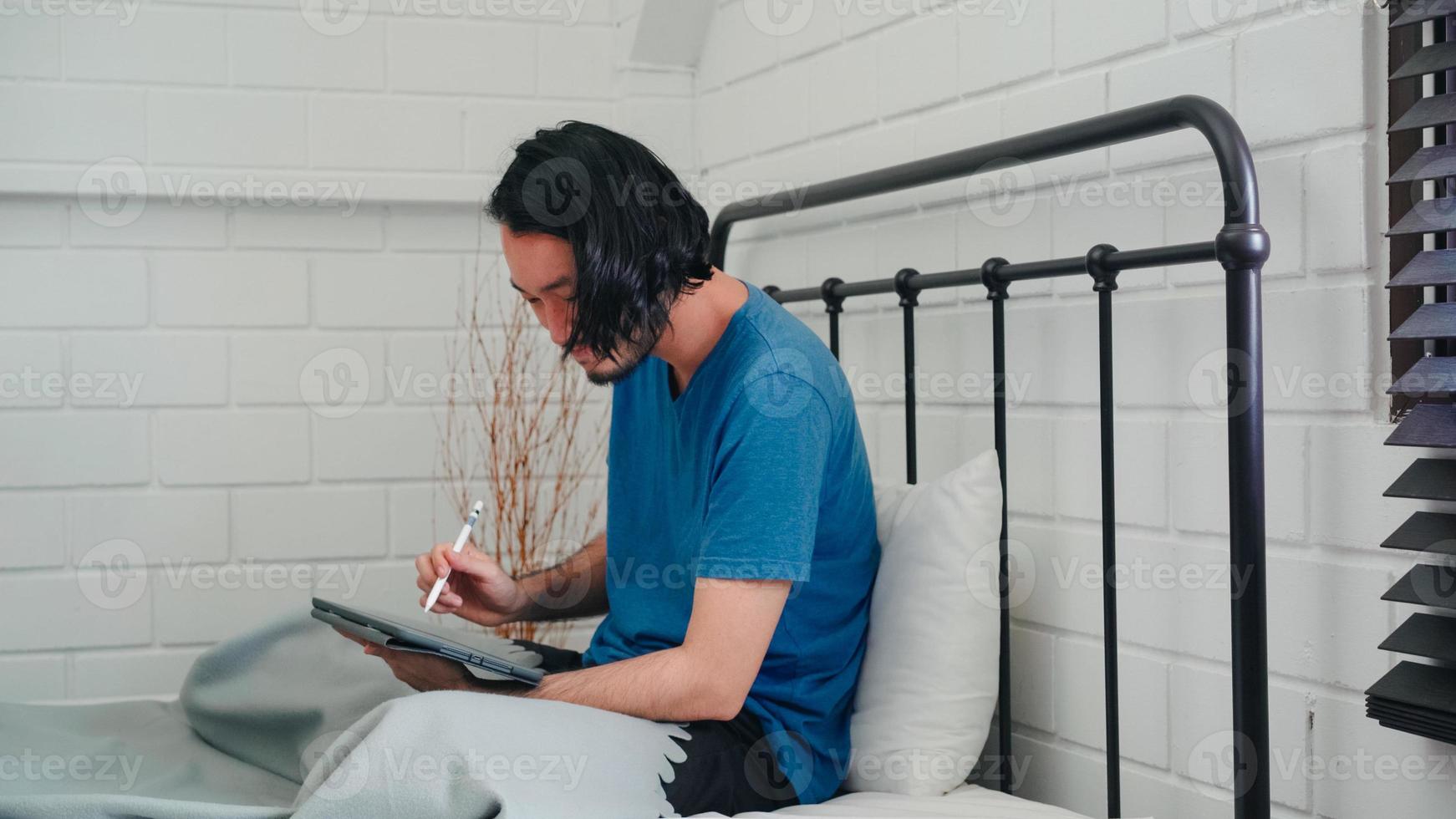 Young Asian man using tablet checking social media feeling happy smiling while lying on bed after wake up in the morning, Attractive male smiling relax in bedroom at home concept. photo