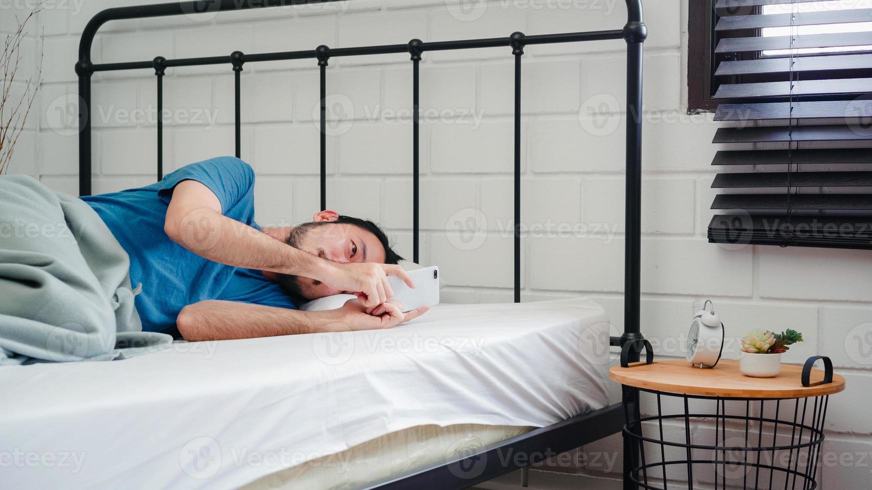 Young Asian man wake up in the morning, Asia male using mobile phone check social media and send message after awake on bed in bedroom at home. Handsome men nap, sleepy relax in modern house concept. photo