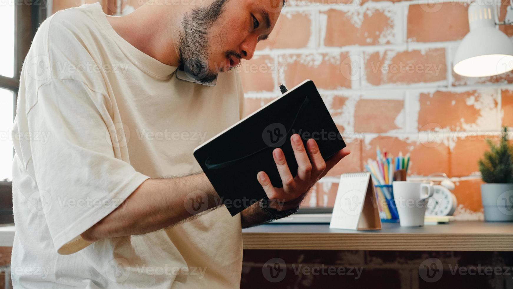 Freelance Asia guy working at his desk in office taking a call on his mobile phone while writing notes on a notepad. Working from home, remotely work, social distancing, quarantine for coronavirus. photo