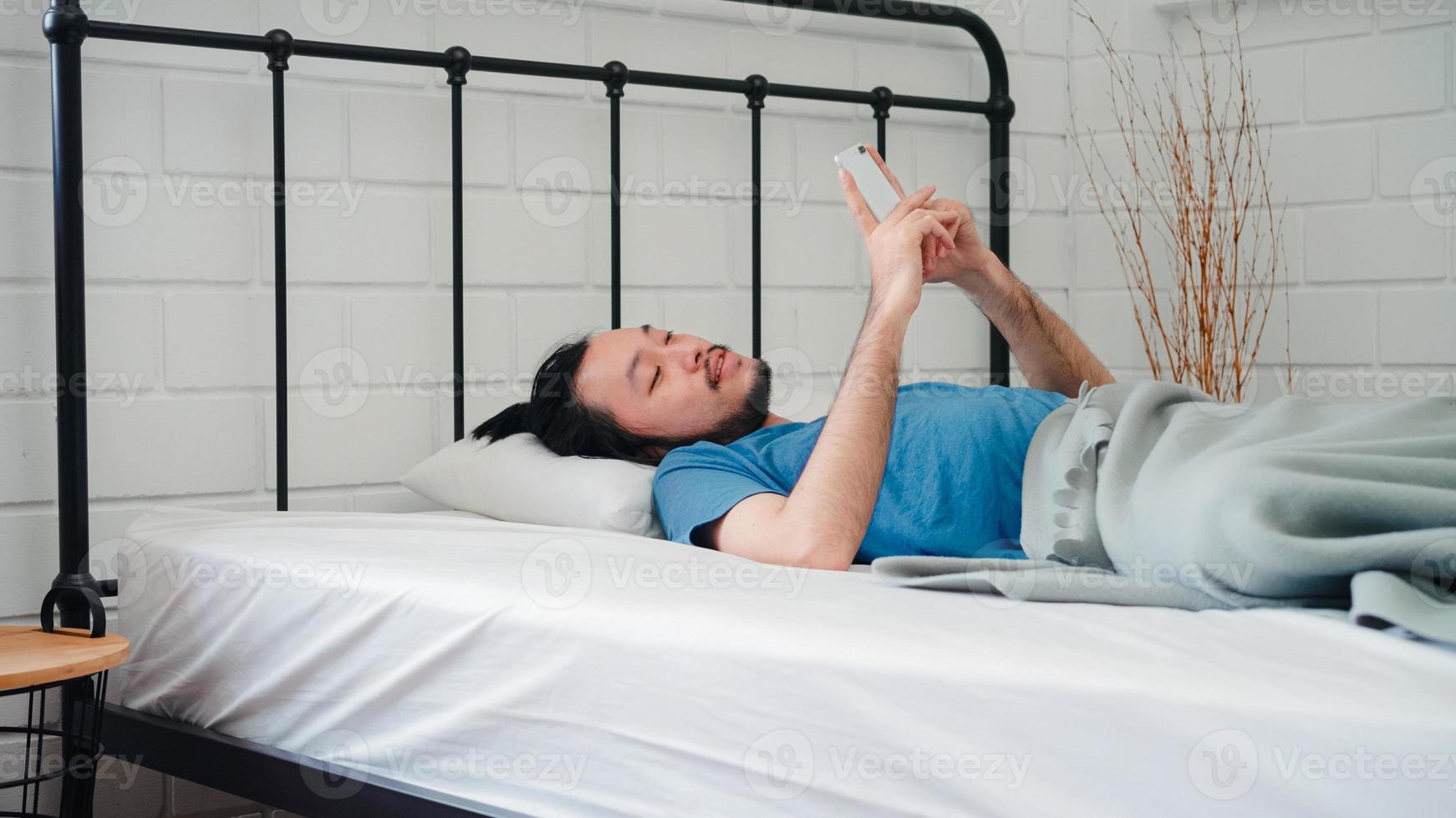 Young Asian man wake up in the morning, Asia male using mobile phone check social media and send message after awake on bed in bedroom at home. Handsome men nap, sleepy relax in modern house concept. photo