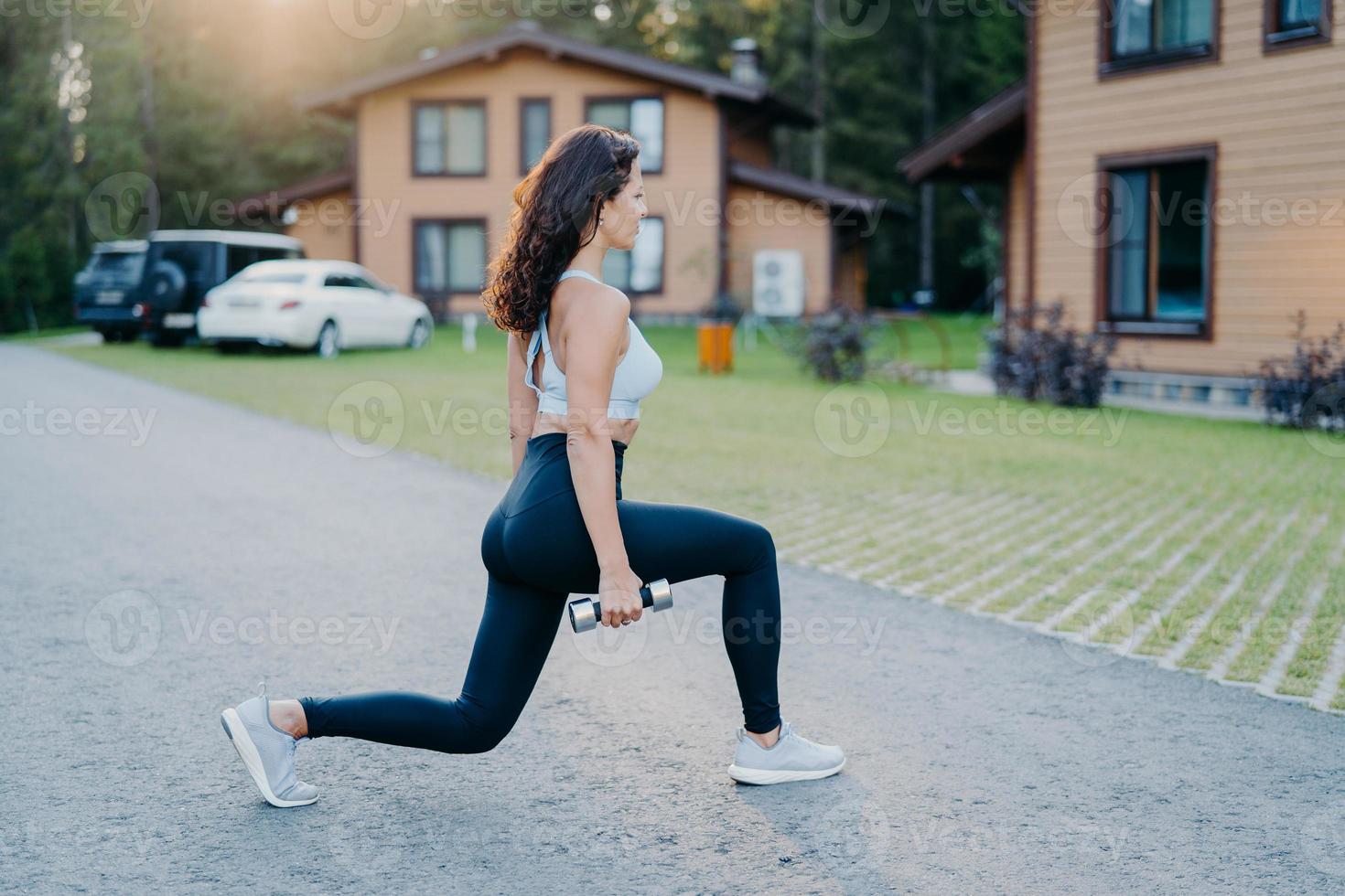 Una foto al aire libre de una mujer saludable en forma vestida con ropa activa hace ejercicios físicos con pesas en trenes al aire libre, poses de bíceps afuera con casas en el fondo. deportista disfruta aeróbic