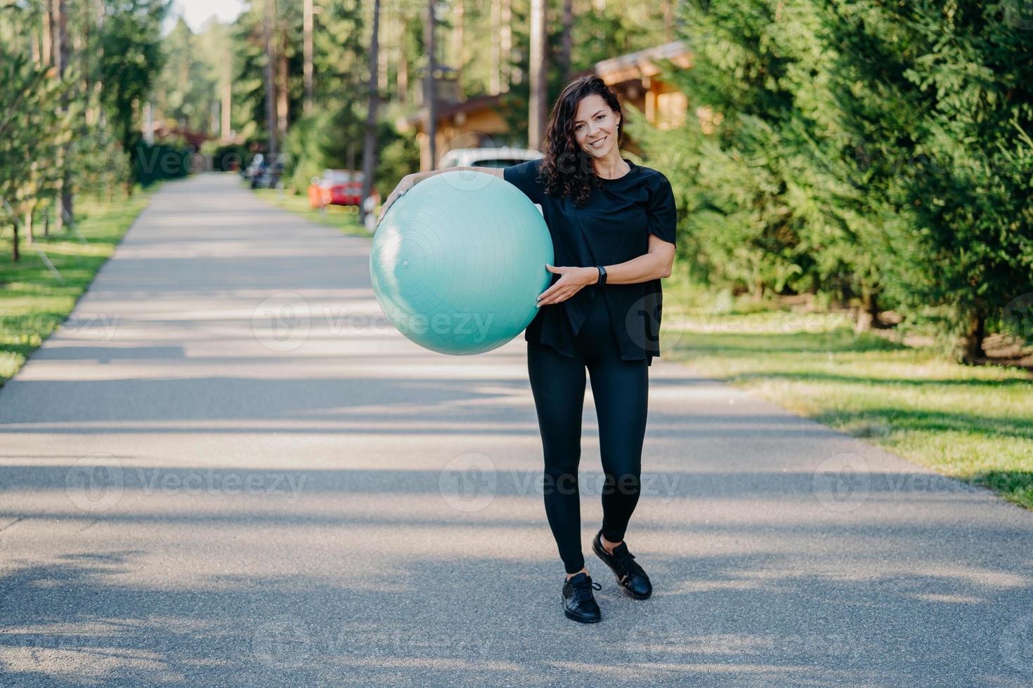 Fitness woman exercises with fitness ball outdoor walks on road near forest has healthy lifestyle dressed in black active wear, has happy expression, stays in good physical shape. Fitness concept photo