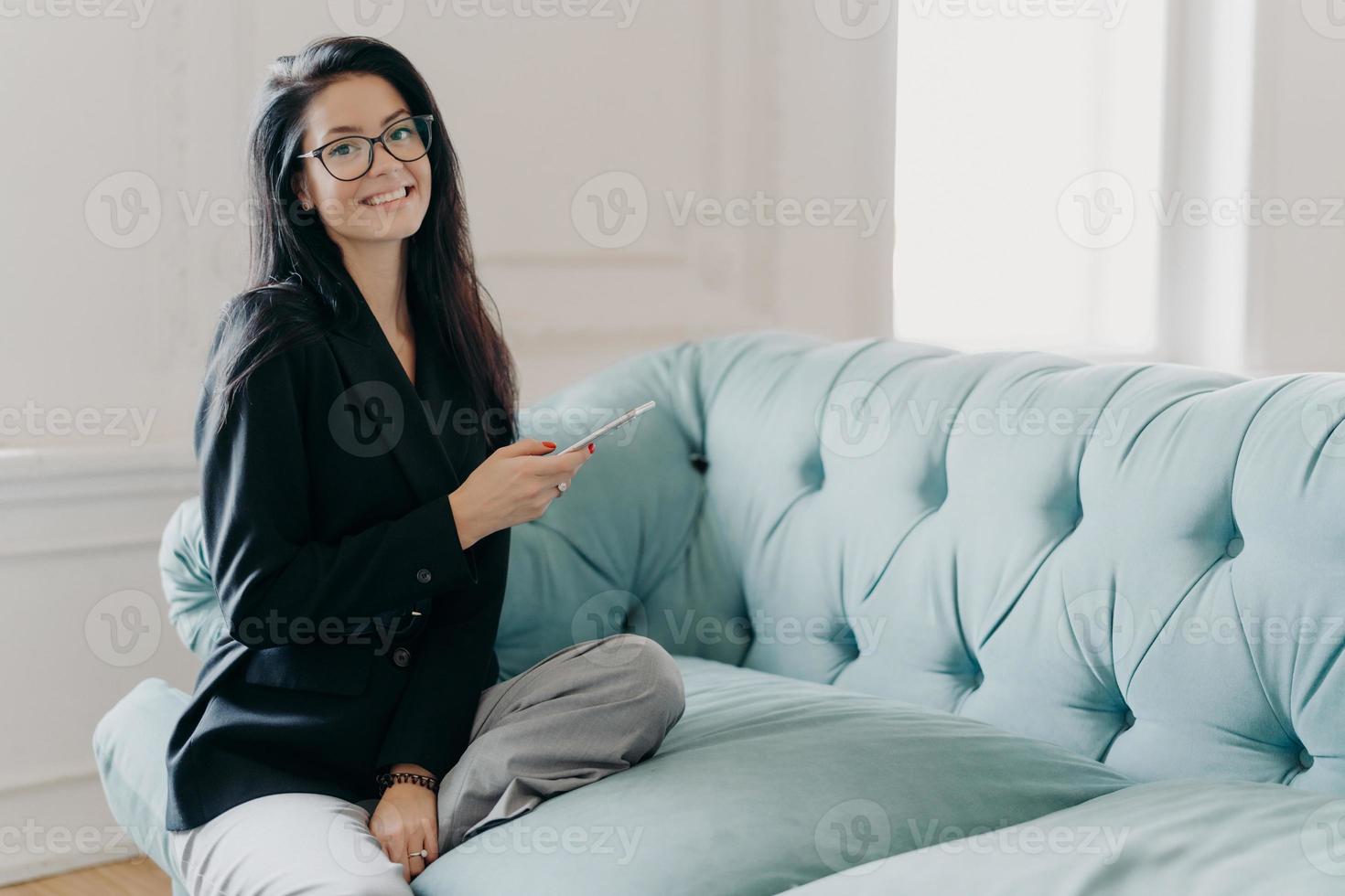 mujer de negocios feliz con ropa oficial, se sienta en un cómodo sofá, usa el teléfono móvil, lee noticias a través de la red, busca información en el celular, va al compañero de teléfono, explora el proyecto en línea foto