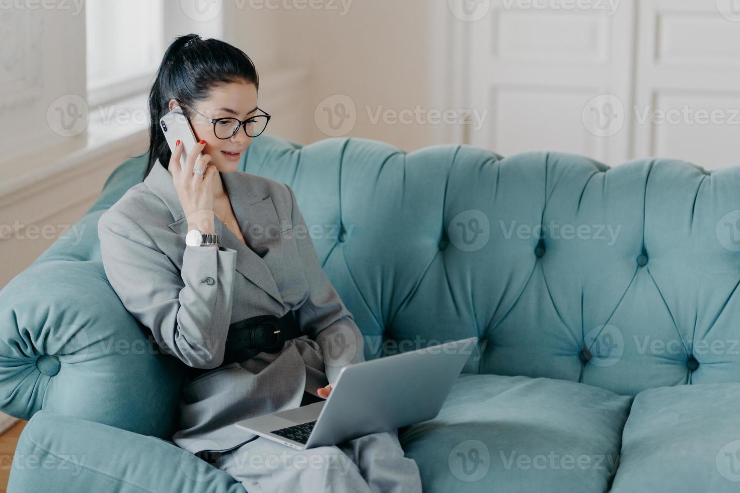 Busy female designer has cell phone conversation, sits in front of opened laptop computer on comfortable sofa, discusses new project. Businesswoman dressed in formal suit works remotely from home photo