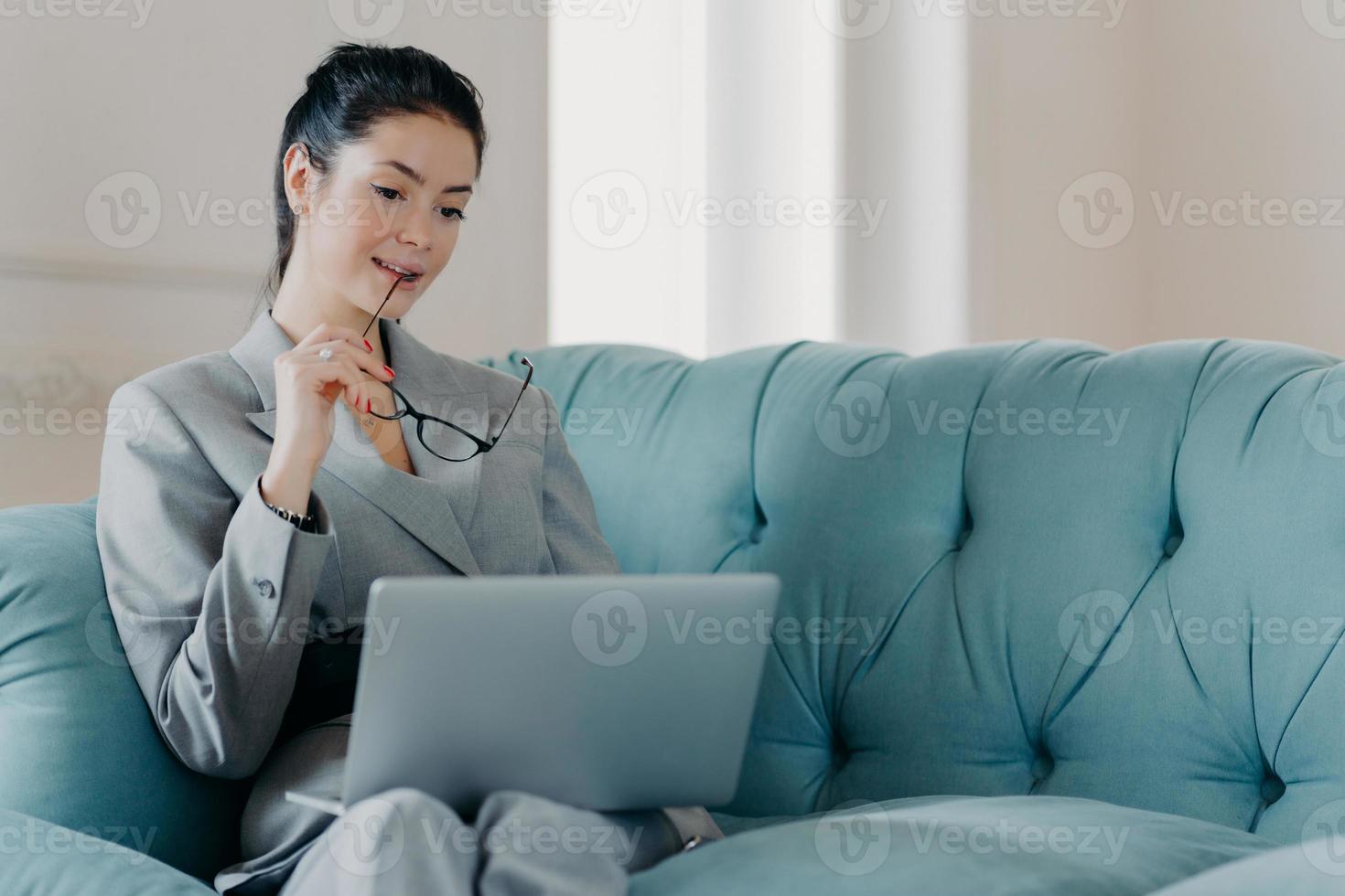 Serious woman entrepreneur takes off glasses, focused in laptop computer screen, analyzes statistics data, looks through financial information, works online, sits on sofa in coworking space. photo