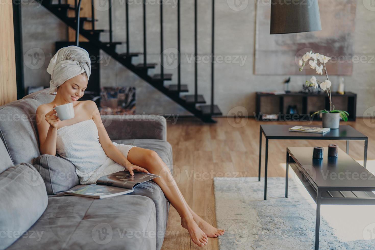 Relaxed woman with slender legs, healthy skin, enjoys drinking tea, sits on comfortable sofa, wrapped in bath towel, poses against cozy home interior. People, beauty, relaxation, lifestyle concept photo