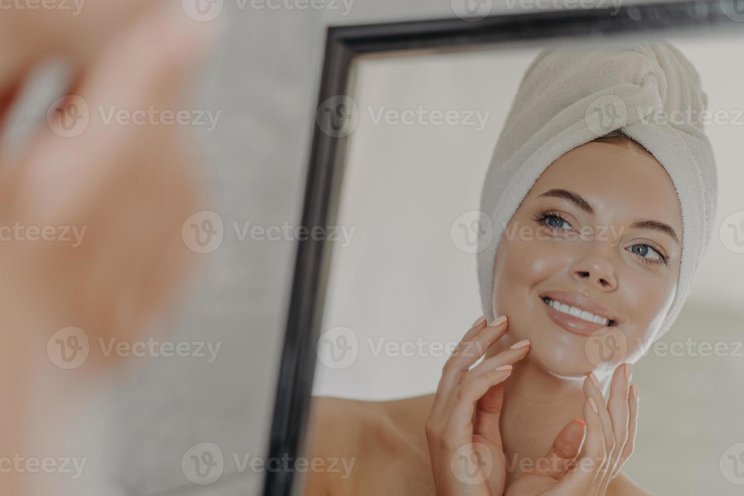 Healthy adorable young woman looks at her reflection in mirror, touches healthy glowing smooth skin, wears minimal makeup, bath towel wrapped on head after taking shower. Beauty routine concept photo