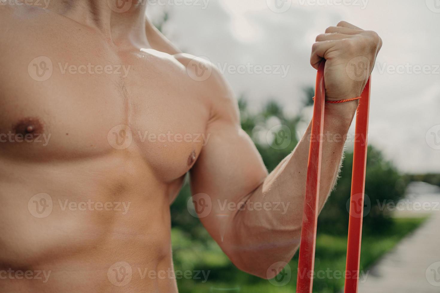 hombre fuerte irreconocible tiene entrenamiento con banda de resistencia, se vuelve más fuerte cada día, muestra un cuerpo bien construido, posa afuera. concepto de deporte, culturismo y estilo de vida saludable. atleta sin rostro foto