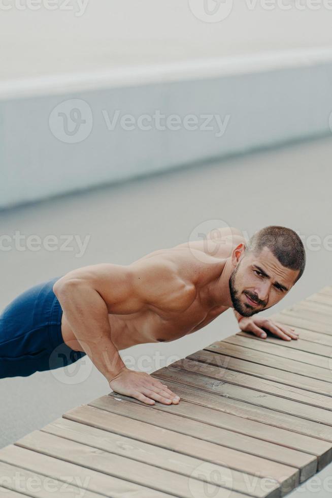 Vertical shot of muscular bearded man stands in plank pose trains chest and arms muscles has hard workout for keeping athletic body shape poses outdoor. Active lifestyle and motivation concept photo